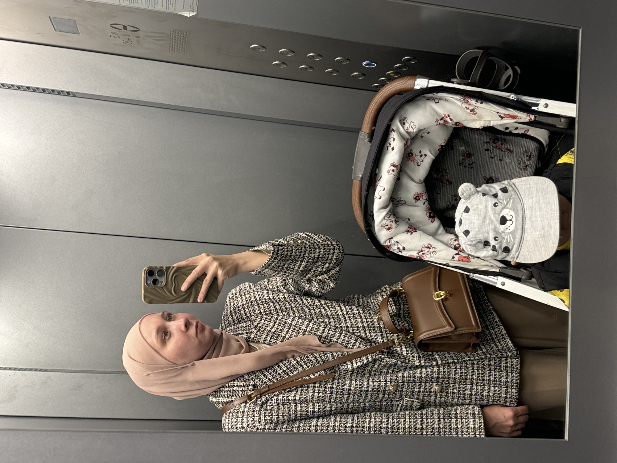 A woman wearing a tan hijab, a tweed jacket, and a brown purse with a strap is taking a mirror selfie in what appears to be an elevator. A baby stroller is positioned next to her. The elevator has a gray metallic interior with numerous buttons. The location is in Chaikovsky, Russia, based on the woman's location history.