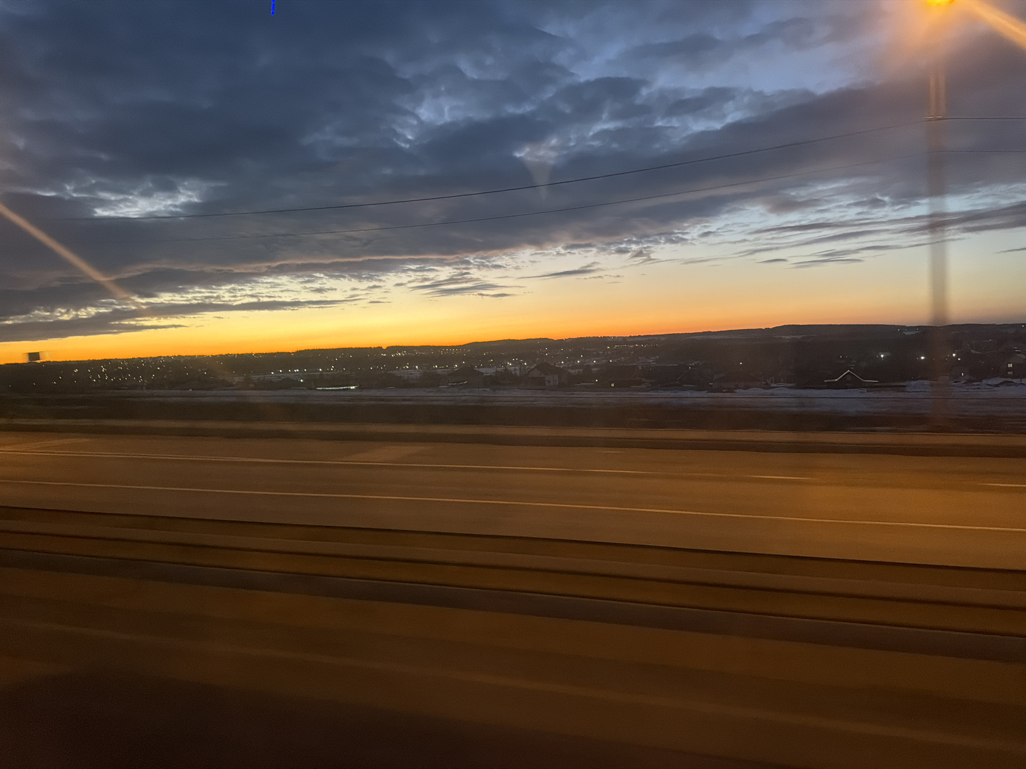 The image depicts an elevated view of a multi-lane roadway at twilight in Kosoturikha, Russia, under a dramatic sky. The upper sky features dark blue-grey altocumulus clouds, transitioning to a bright orange and yellow horizon, indicating either sunrise or sunset. Horizontal utility lines traverse the mid-sky. A distinct diagonal light streak, consistent with a sunbeam or lens flare, originates from the upper-left quadrant, while a prominent vertical light streak from an artificial light source is visible on the far right. In the foreground, a multi-lane asphalt road, illuminated by an orange-brown artificial glow, extends horizontally across the lower frame, displaying white lane markings for at least three lanes. No vehicles or pedestrians are present on the roadway. Beyond the road, an uneven terrain with visible patches of snow and ice leads to a sprawling settlement identified as part of Kosoturikha. This area is characterized by numerous scattered artificial lights emanating from buildings and infrastructure against a dark, undulating distant horizon.
