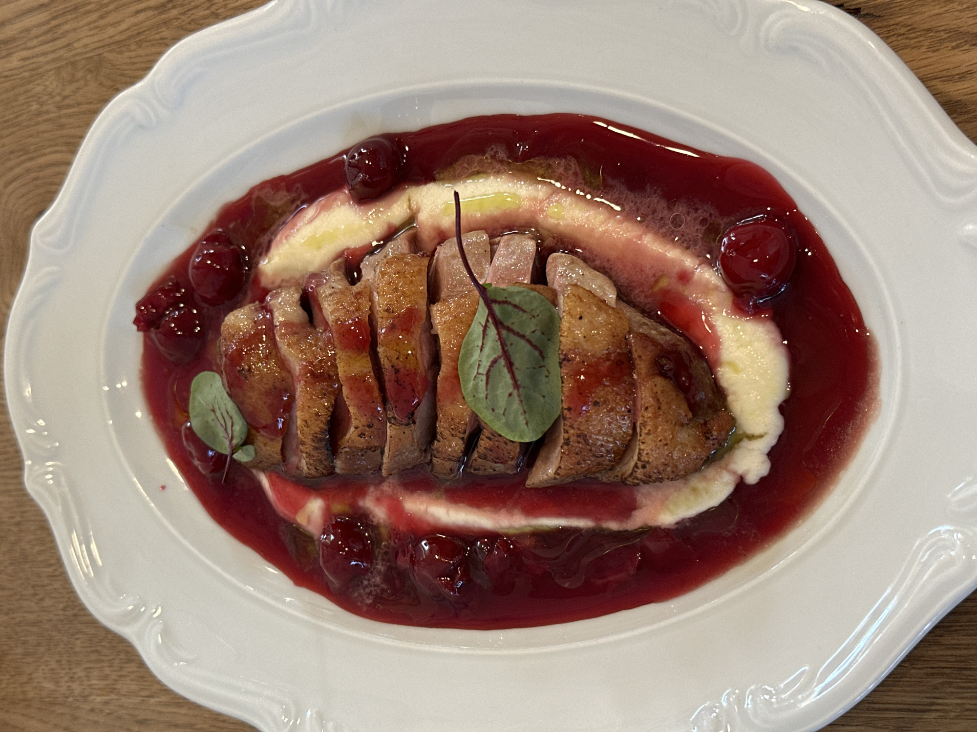 A top-down, eye-level close-up shot shows a plate of food. The food consists of sliced duck breast resting on a bed of cream-colored purée and surrounded by a dark red cherry sauce with whole cherries. Two small green leaves garnish the dish. The plate is white with a decorative edge and sits on a light brown wooden surface. The location is Unknown, Unknown.