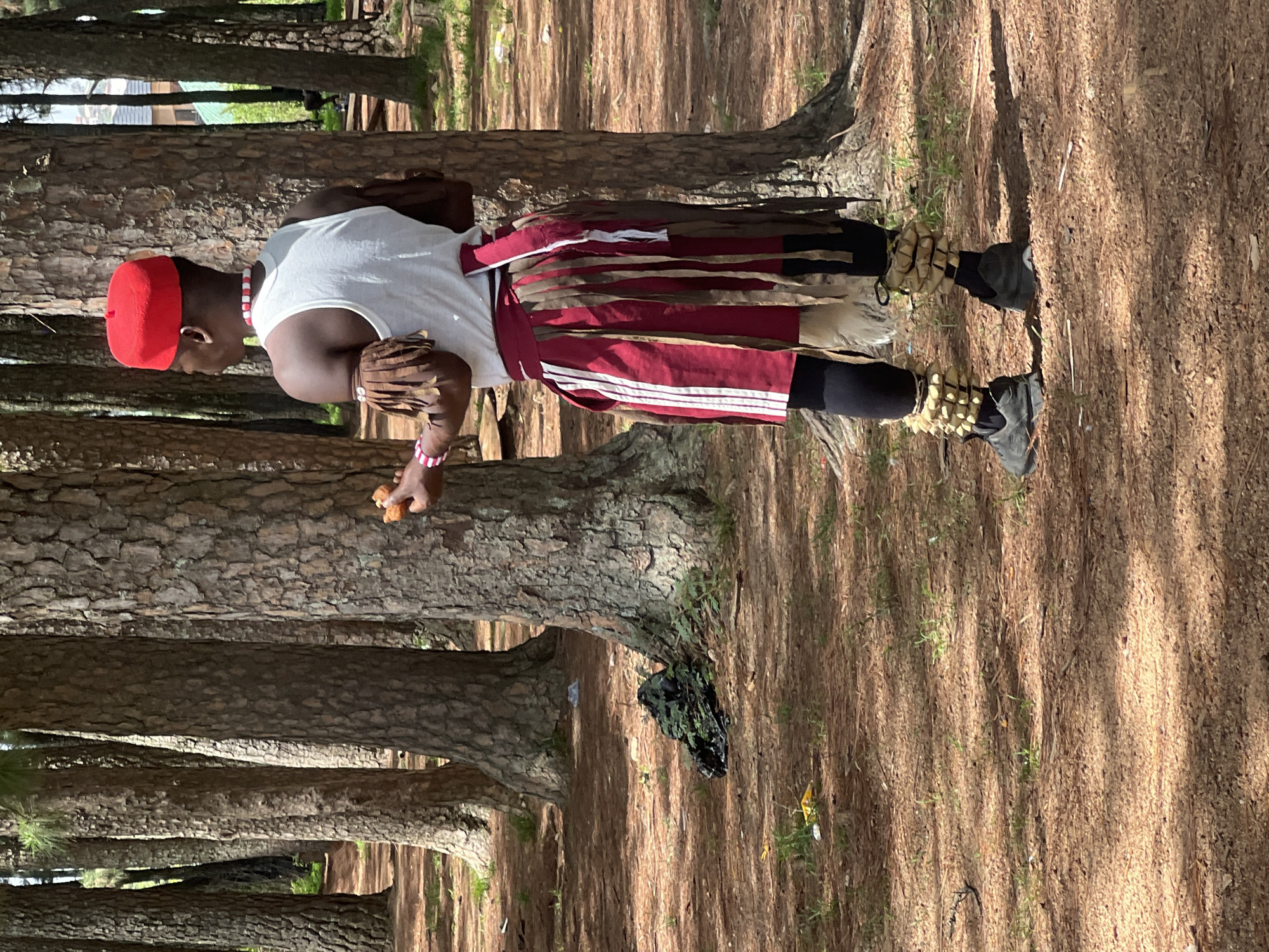 In a sunlit, pine forest setting near Jos, Nigeria, a man stands in traditional Igbo attire. He wears a red cap, a white tank top, a red and white striped wrapper, black leggings, and footwear adorned with bone-like embellishments. He is holding a small, unidentifiable object in his hand. The ground is covered with pine needles and the vertical trees cast shadows across the scene.