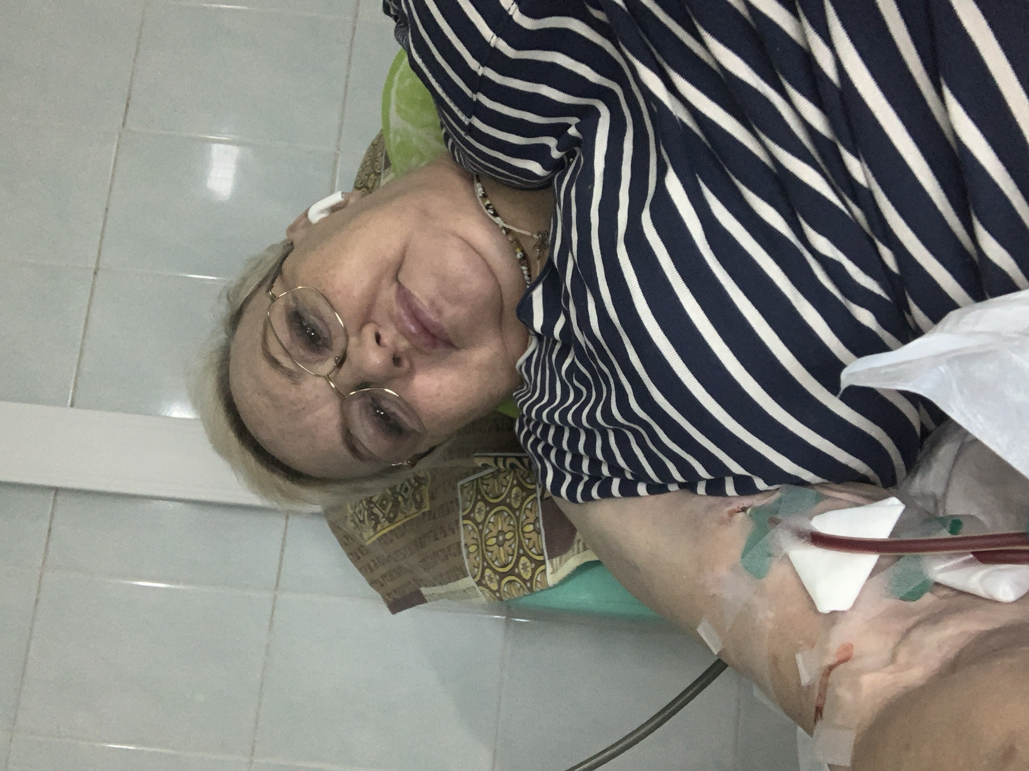 A woman lies in a bed in a hospital room. She wears gold-rimmed glasses, a striped top, and an earbud in her left ear. An IV line is inserted into her left arm. The room has tiled walls. There is no information available to determine the location to be Chaikovsky, Russia.