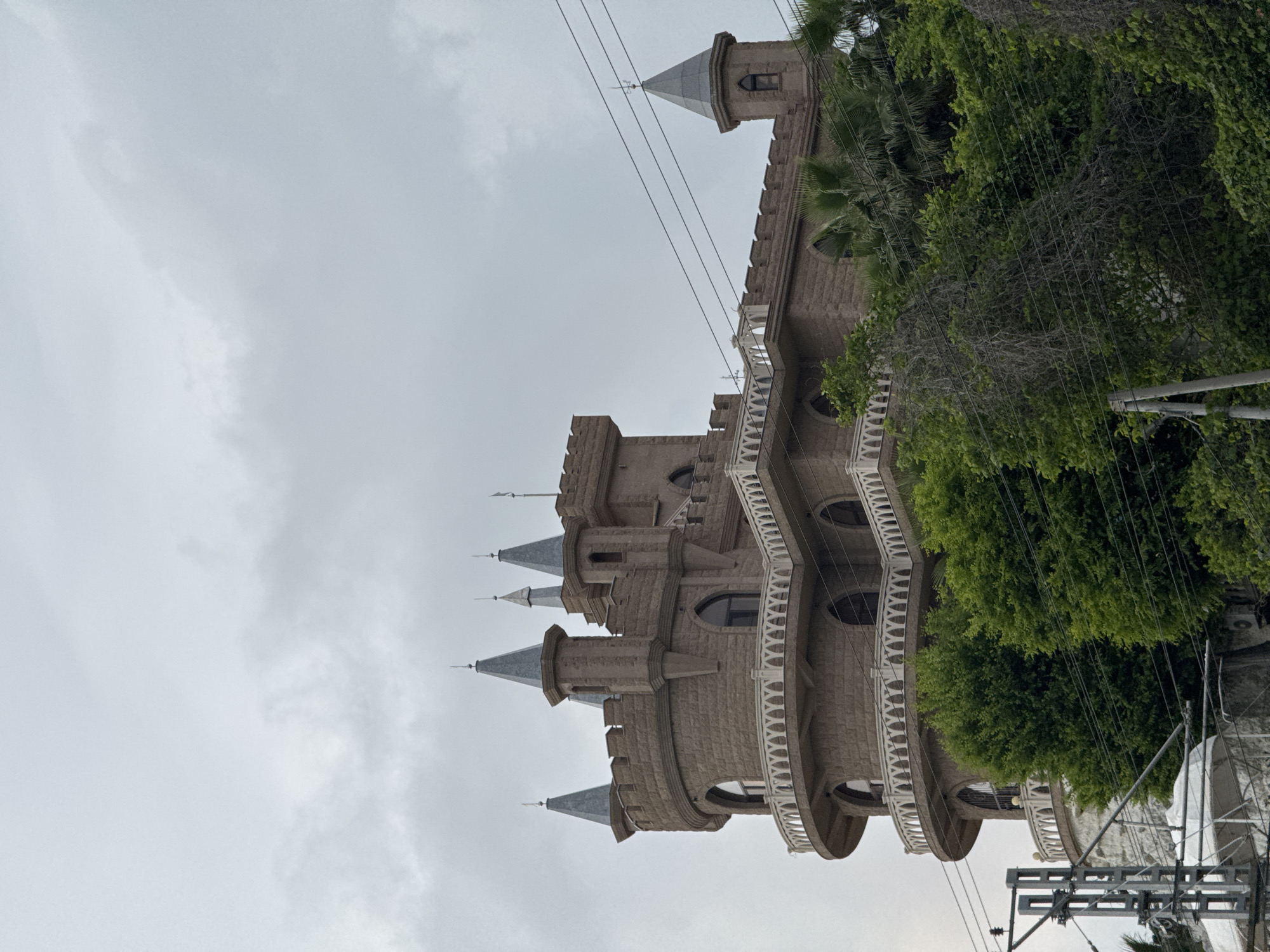 The image shows a castle-like building with multiple towers and turrets against a cloudy sky. The building appears to be constructed from stone and features rounded balconies with white railings. The surrounding area includes trees and utility poles with power lines. Based on prior knowledge, this structure is likely located in Sochi, Russia, based on its architectural style and common images of the city. No people are visible in the image.