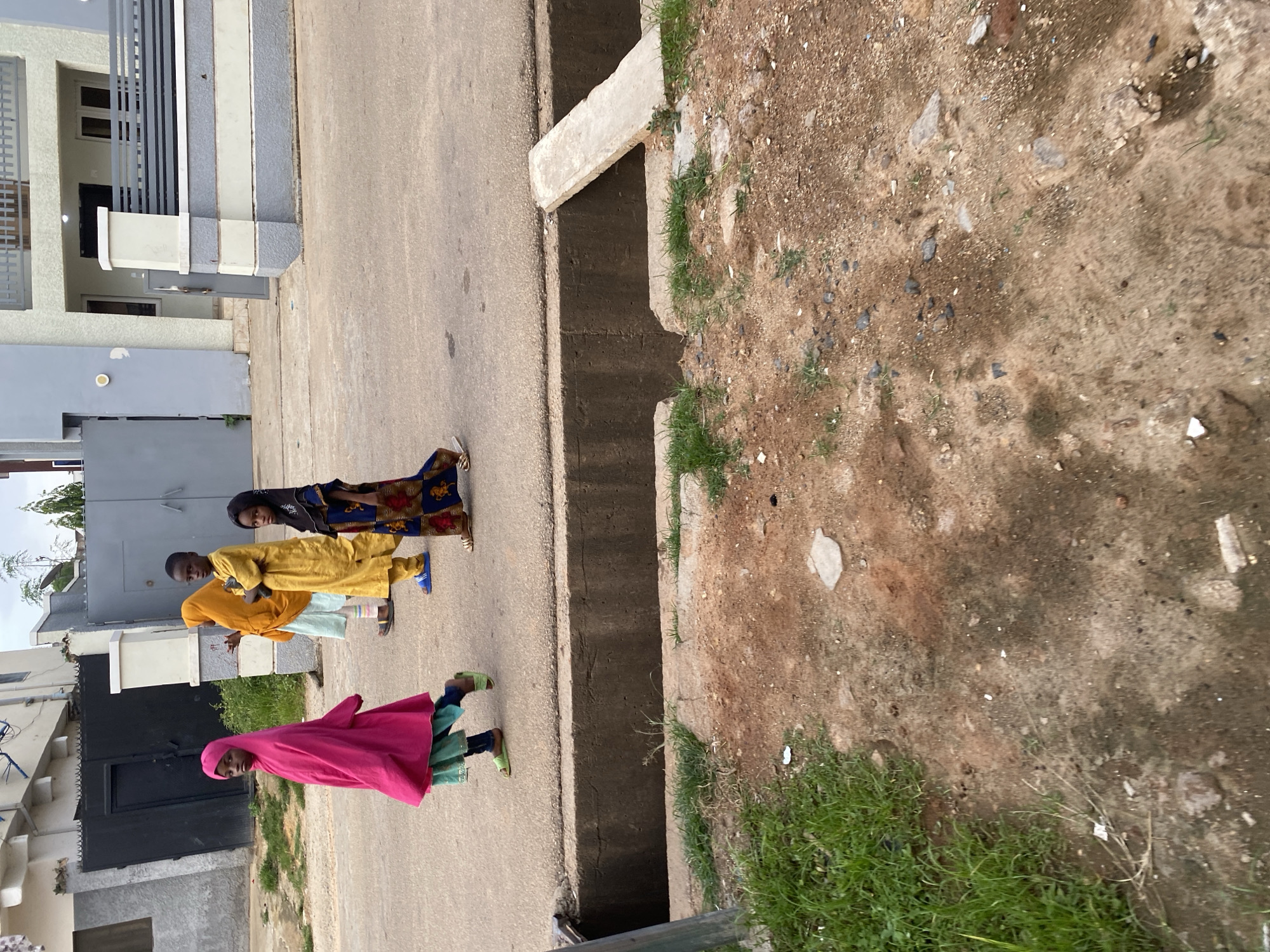Four children are walking on a paved road in front of a residential building in Jos, Nigeria. The child on the far left is wearing a bright pink hijab and a green dress. Next to her is a child wearing a yellow hijab and a yellow garment. To their right, a child in a blue and gold patterned dress with a black headscarf is walking. In the foreground, a concrete drainage ditch runs along the side of the road. The area appears to be a residential street with buildings and a black metal gate visible in the background. The ground on the side of the road is a mix of dirt and grass.