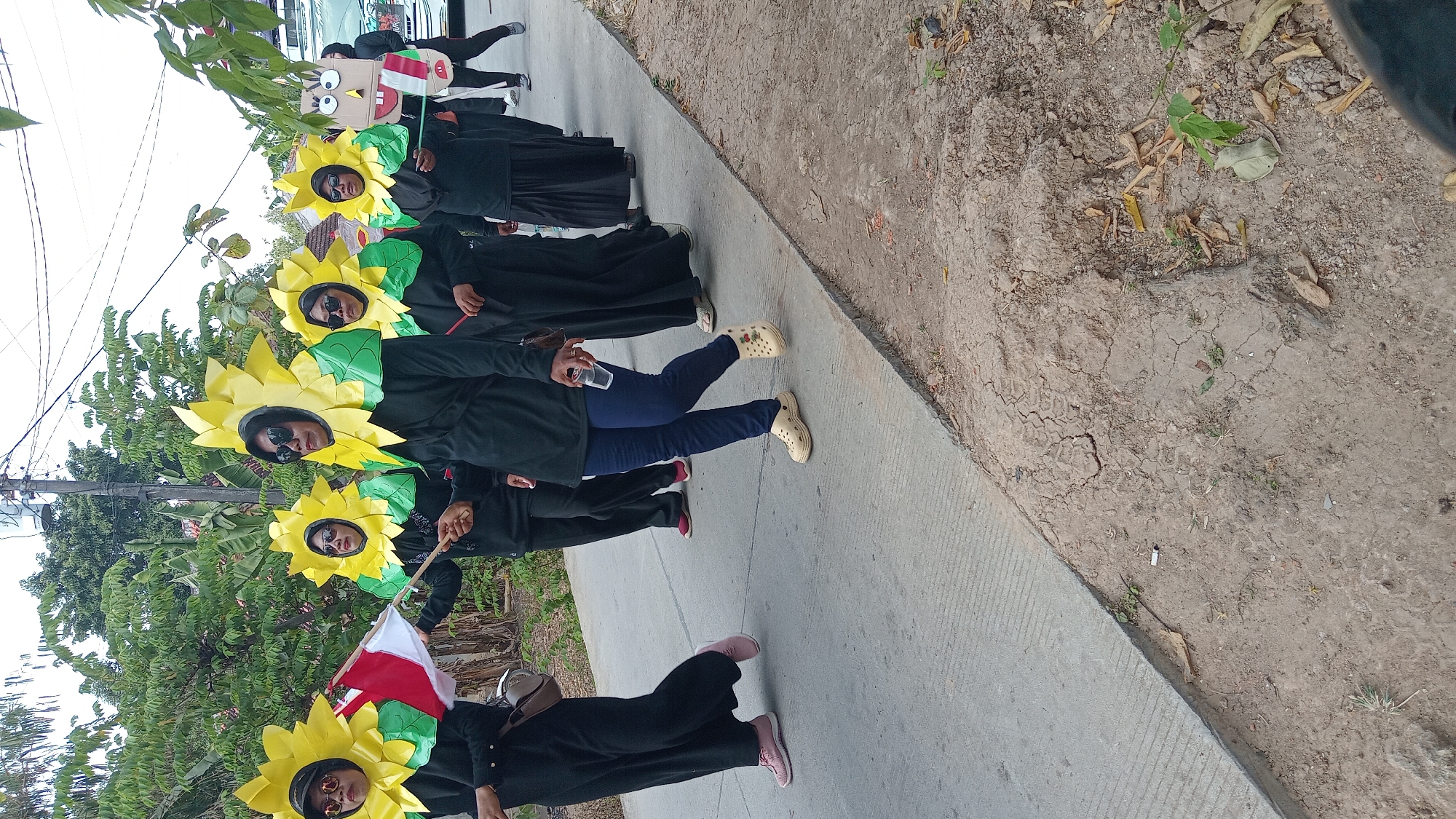 In Belor, Indonesia, a group of individuals dressed in sunflower costumes walks along a paved road. The costumes feature large yellow petals and green leaves around the wearers' heads. The individuals are wearing black clothing, and some are wearing sunglasses. One person in the front carries a small Indonesian flag. Behind the group, a vehicle can be seen. The environment appears to be outdoors, with trees and vegetation visible in the background, suggesting a street or roadside in a residential or semi-rural area. The ground is a mixture of concrete and dry, cracked earth.