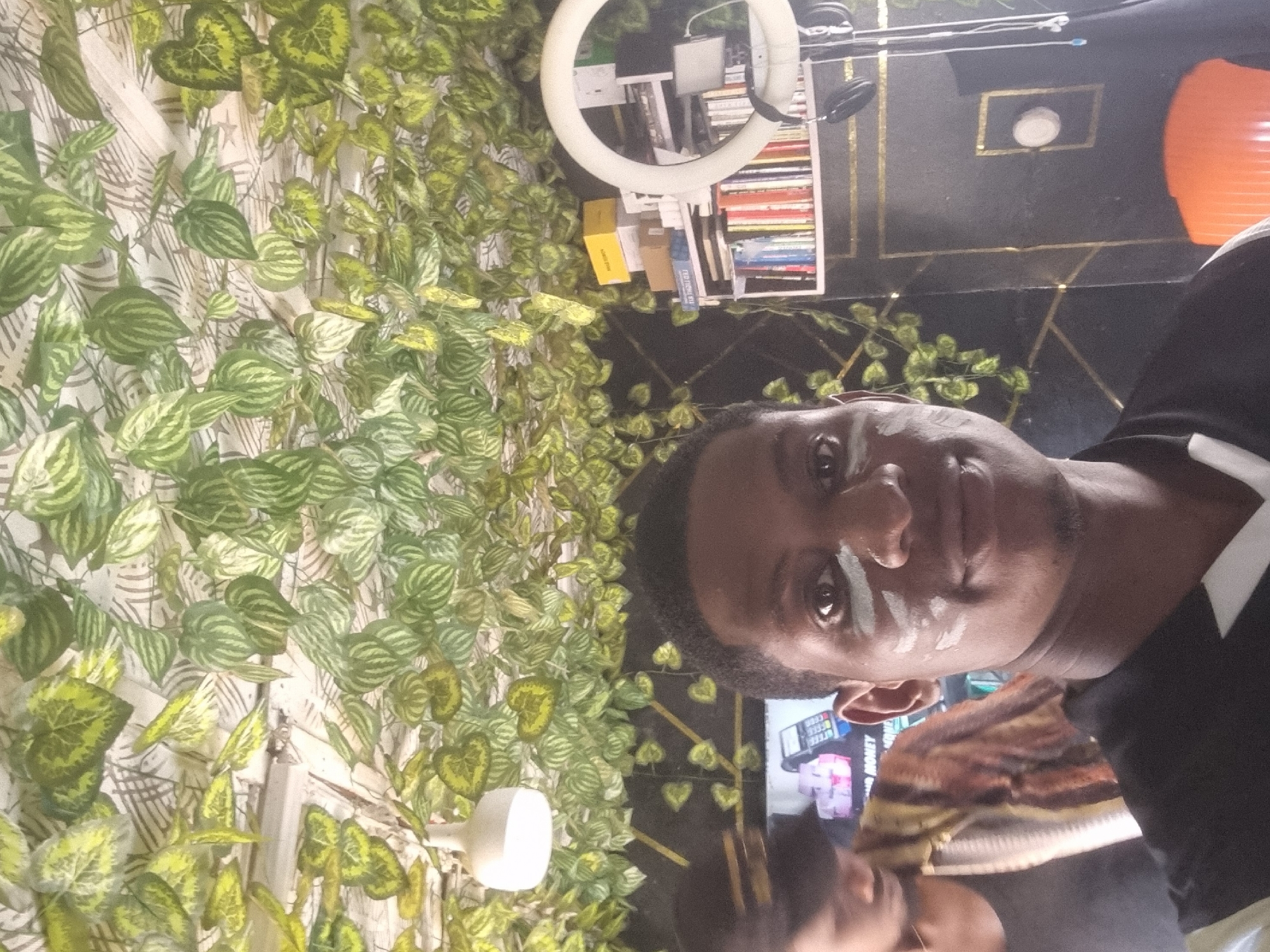 A man with short, dark hair and light streaks of white cream on his face is standing indoors. He is wearing a black shirt with a white collar. The backdrop is decorated with artificial green leaves and vines covering the wall. Behind him, there is a bookshelf with books and a ring light. The ceiling is dark with geometric gold outlines. In the background, there is a television screen showing an advertisement. The location is potentially in a room or studio in the city of Onitsha, Nigeria.