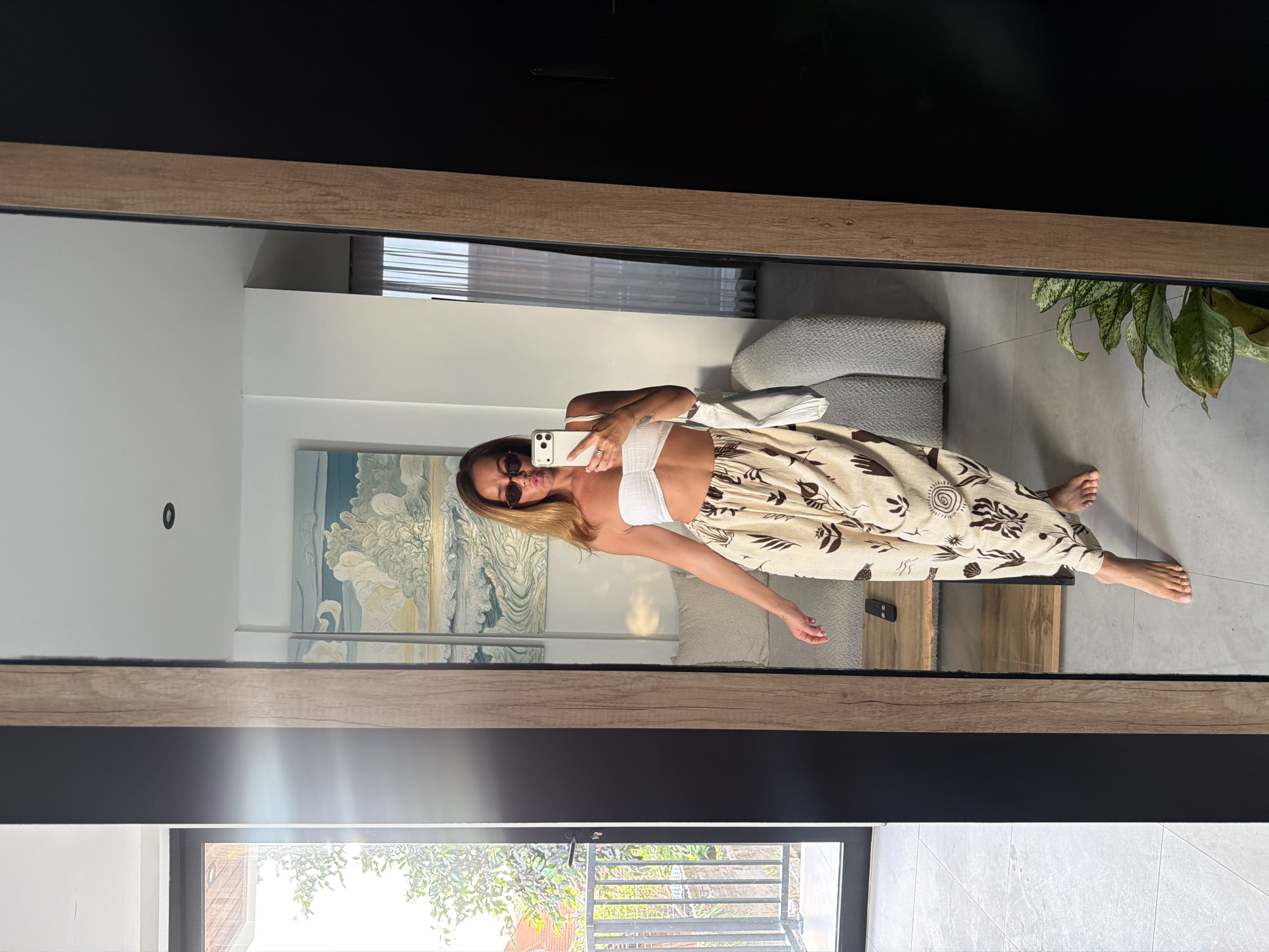 A full-shot, mirror reflection photograph captures a woman standing in a room, holding a smartphone to take a selfie. She is positioned to the left of the frame, facing the mirror. The woman has long, blonde hair and is wearing a white bandeau top and light-colored, loose-fitting pants with a dark brown abstract pattern. She is wearing sunglasses and has her lips pursed. On her left side, she is holding a white bag.
The environment appears to be a modern living space, reflected in a large, dark-framed mirror that dominates the right side of the image. To the left of the mirror, a doorway opens to an outdoor balcony with a railing and lush green foliage visible beyond. Inside the room, a light-colored couch is visible behind the woman. A wooden shelf unit with a black remote control on it is situated in the mid-ground of the reflection. A large piece of artwork depicting a seascape with clouds hangs on the wall above the couch. A potted plant with green and white leaves is visible in the lower right corner of the reflected scene. The floor is tiled in a light gray color. The overall lighting suggests daytime with natural light entering from the left.