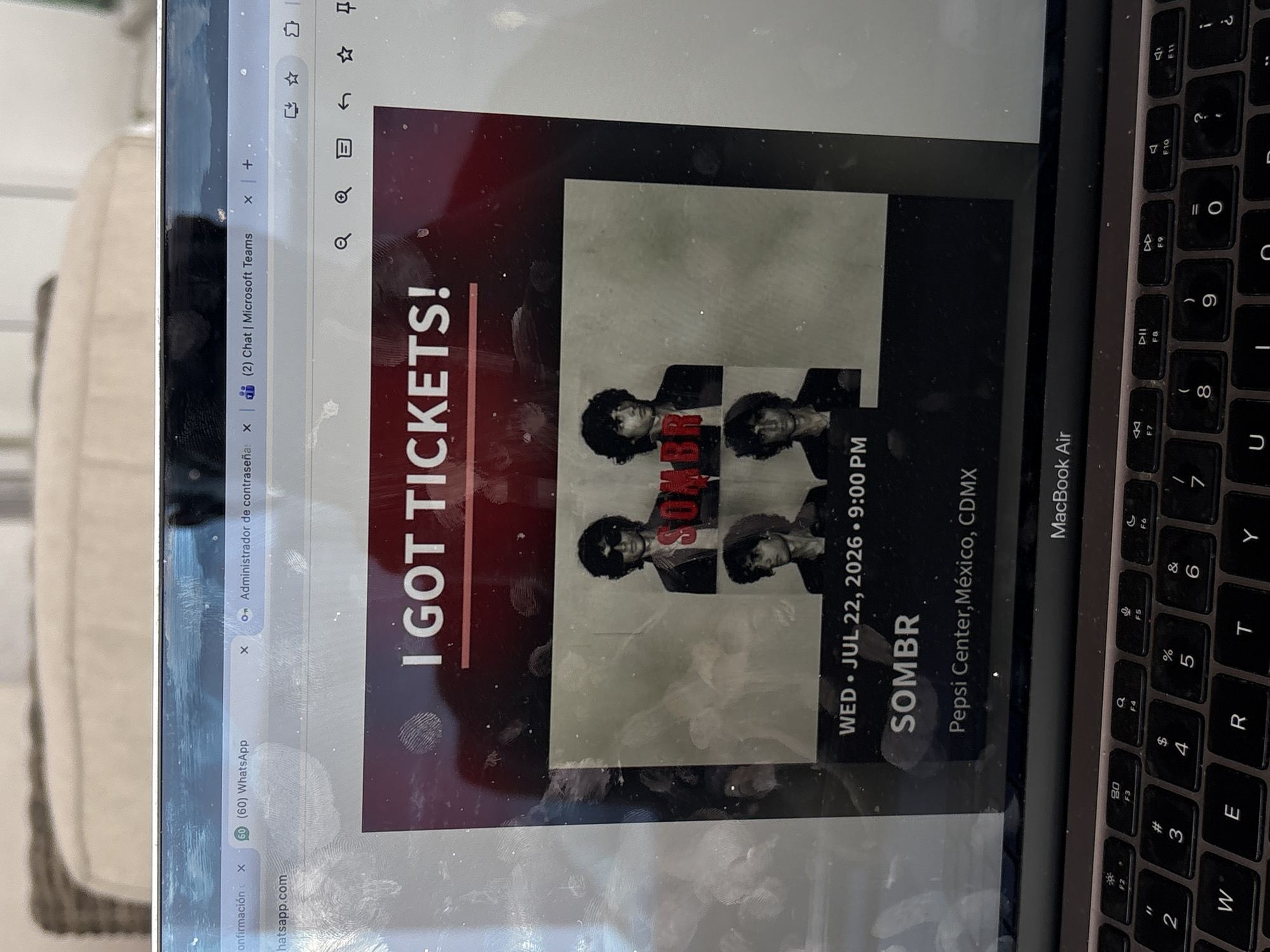 Here is a description of the image:
The image shows a close-up of a laptop screen displaying a concert ticket. The ticket announcement says "I GOT TICKETS!" The band performing is "SOMBR", and the concert will be held on Wednesday, July 22, 2026, at 9:00 PM at the Pepsi Center in Mexico City, Mexico. The laptop is a MacBook Air, and the keyboard is visible in the lower right corner of the image. The screen is smudged with fingerprints. Several browser tabs are open at the top of the screen, including WhatsApp, a password manager, and Microsoft Teams. Part of an upholstered piece of furniture is visible to the left.