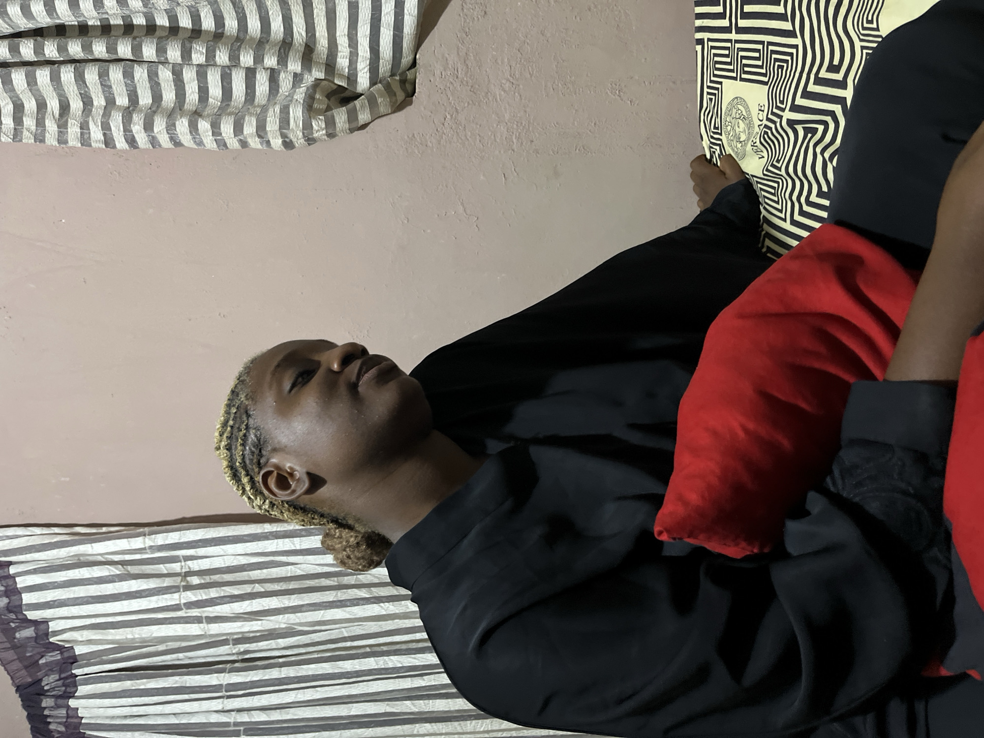 The image shows a young Black woman with blonde braided hair, styled in cornrows and a bun, seated on a bed. She is wearing a black abaya or robe and black pants. A red pillow is propped against her chest, and her left hand rests on a patterned sheet with a Versace logo. The background features a light pink wall and two striped curtains, one on the left with vertical stripes and the other on the right with horizontal stripes. The setting appears to be a bedroom, possibly in Rafin Par, Nigeria, given the context provided. The lighting is dim, creating shadows on the wall and highlighting the texture of the paint. The woman is looking to her right with a slight smile.