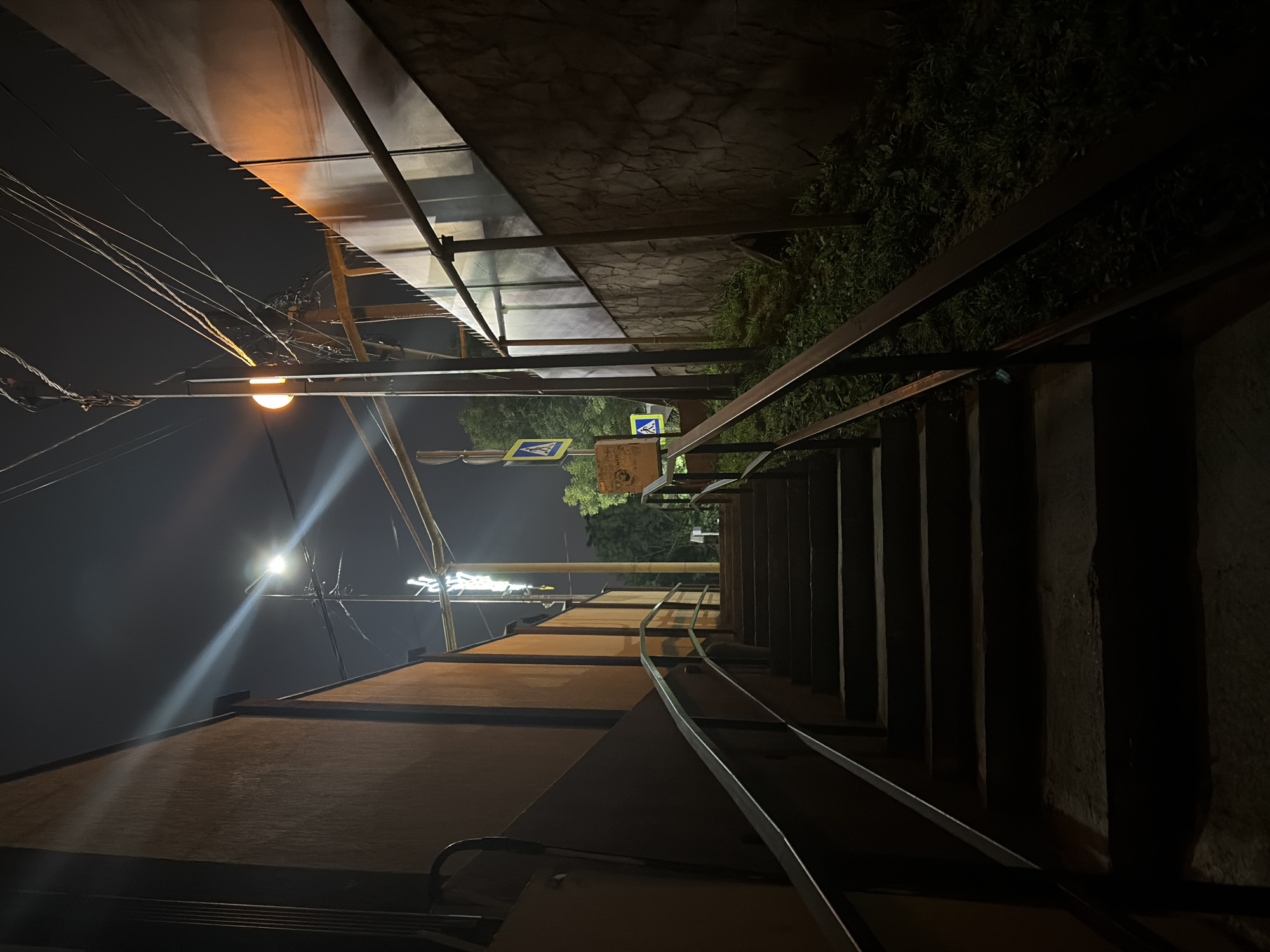 The image depicts an outdoor staircase at night in what is stated to be Sochi, Russia. The staircase is made of concrete with metal railings on both sides, and it ascends into darkness. The surroundings are dimly lit by streetlights and what appear to be neon signs, casting long shadows. To the left of the stairs, a textured orange wall runs parallel, while to the right, a stone-clad retaining wall is visible, overgrown with grass. Overhead, a network of power lines crisscrosses the sky. A yellow pedestrian crossing sign is mounted on a pole further up the stairs on the right side. No people are present in the image.