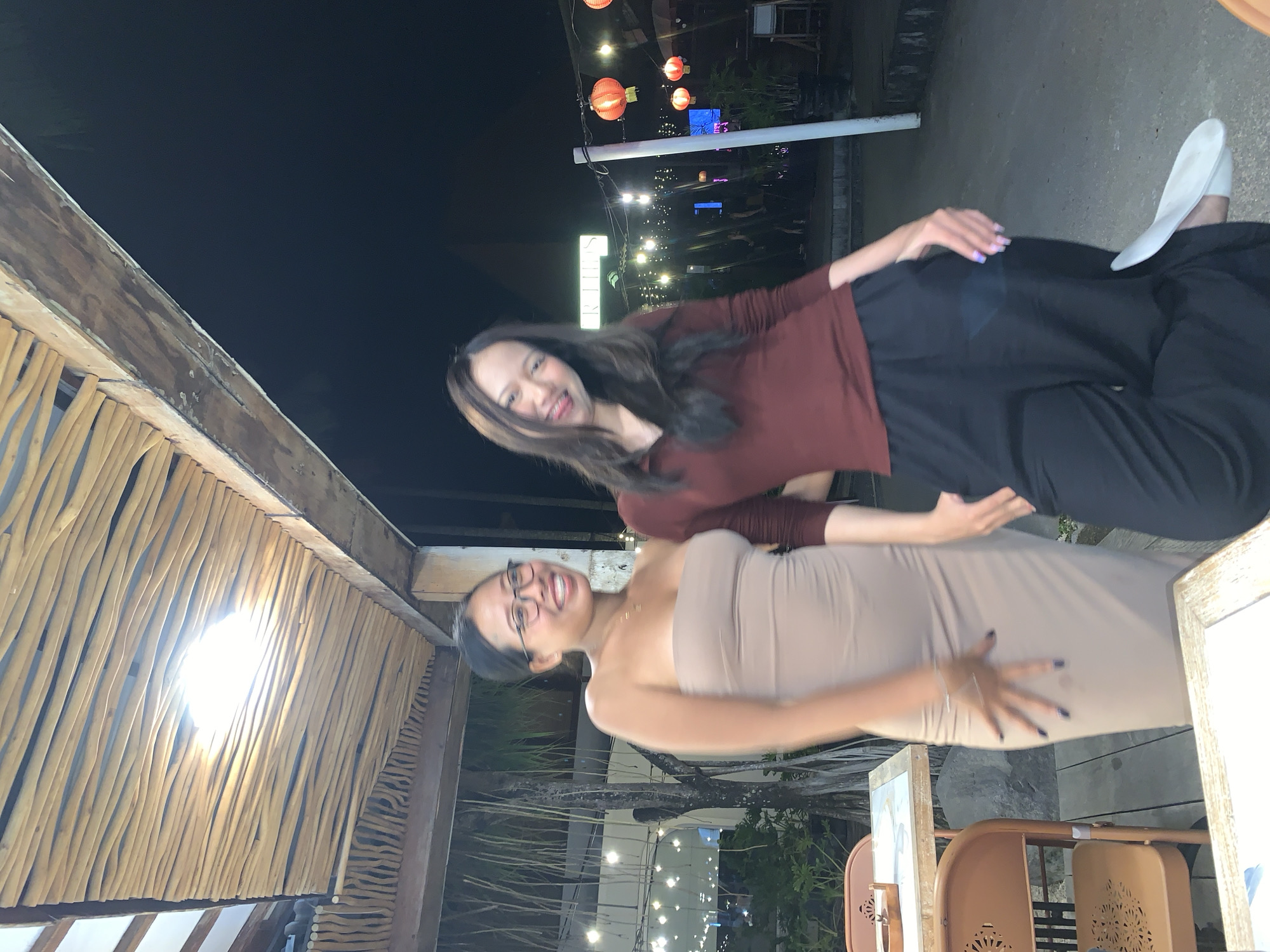 Two women are posing for a picture outdoors at night. One woman is wearing glasses and a tan dress, while the other is wearing a maroon top and black pants. The location appears to be a restaurant or outdoor venue, with lanterns and string lights visible in the background. The photo was taken in North Kuta, Indonesia. A wooden structure with thatched roofing is visible in the upper left.