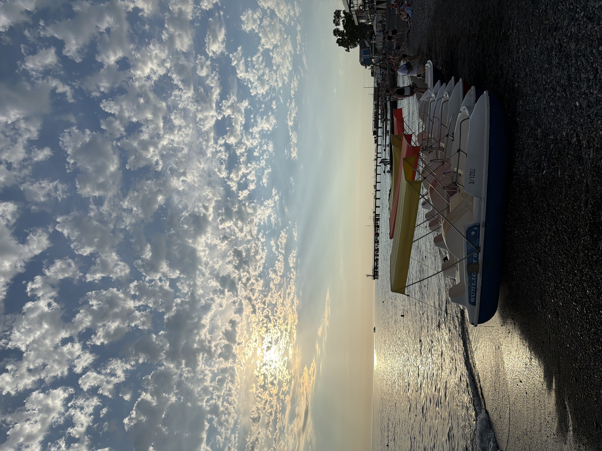 This media file captures a scenic view of a beach in Sochi, Russia. The image is oriented vertically, with the sky occupying the left side and the beach and water on the right. The sky is filled with a dramatic display of cumulus clouds, illuminated by the sun which is partially obscured, suggesting it is either sunrise or sunset. The light from the sun creates a warm glow on the horizon and casts a shimmering reflection on the calm water.
Along the shoreline, a row of white pedal boats with colorful canopies (yellow and red) are lined up on the pebble beach. One of the boats has text visible: "SUNLIGHT T5" and "3/1202". A few people are visible in the background near the boats and on a pier extending into the sea. They appear to be enjoying the beach environment. The overall mood is serene and picturesque, characteristic of a pleasant day at the seaside. The presence of the boats suggests a recreational setting where visitors can rent them for water activities. The time of day is clearly either dawn or dusk due to the low angle of the sun and its soft, diffused light. The weather appears clear and calm, with no indication of rain or strong winds.