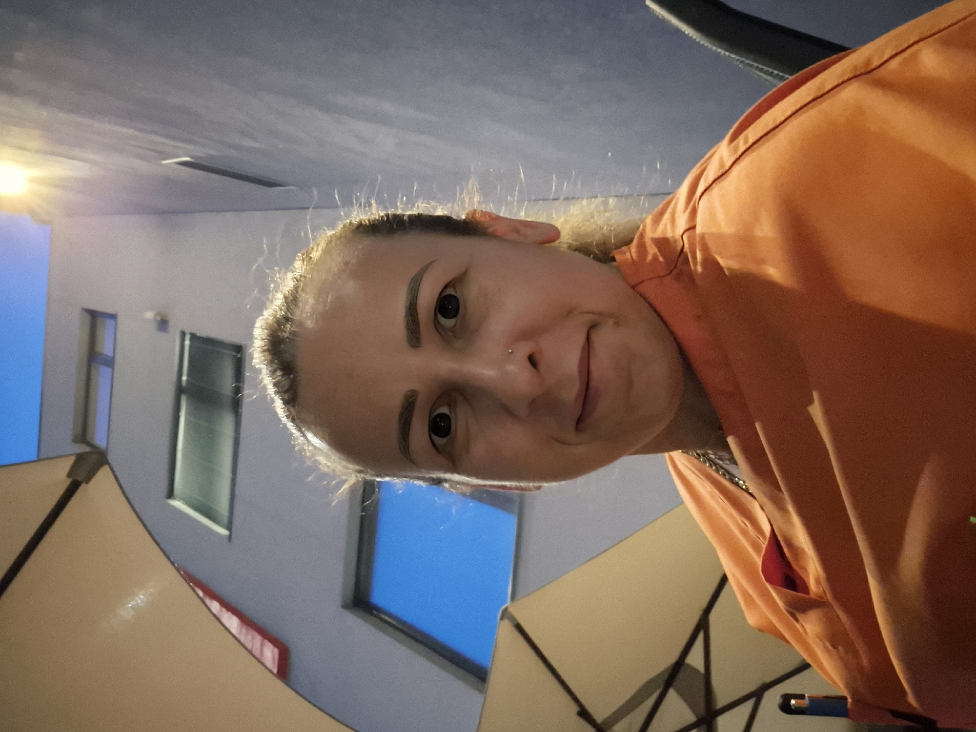 A medium close-up portrait, when correctly oriented, captures a woman looking directly at the camera with a gentle, slightly amused expression. She has light-colored hair, pulled back, with wisps framing her face, dark eyebrows, and a small nose piercing on her right nostril. She is wearing a bright orange top or jacket; a silver chain is subtly visible around her neck.
The setting appears to be an outdoor area, likely a covered patio or terrace. Above the woman, a textured, light grey overhead structure, possibly a ceiling or canopy, is visible. In the background, a building with light-colored walls features several rectangular windows, some of which reveal a bright blue sky, suggesting twilight or early evening. Partially visible in the foreground and mid-ground are sections of cream-colored umbrellas or awnings. A bright, localized light source, possibly artificial, illuminates the scene from the upper left, contributing to the dim ambient lighting. There is no visible text.
