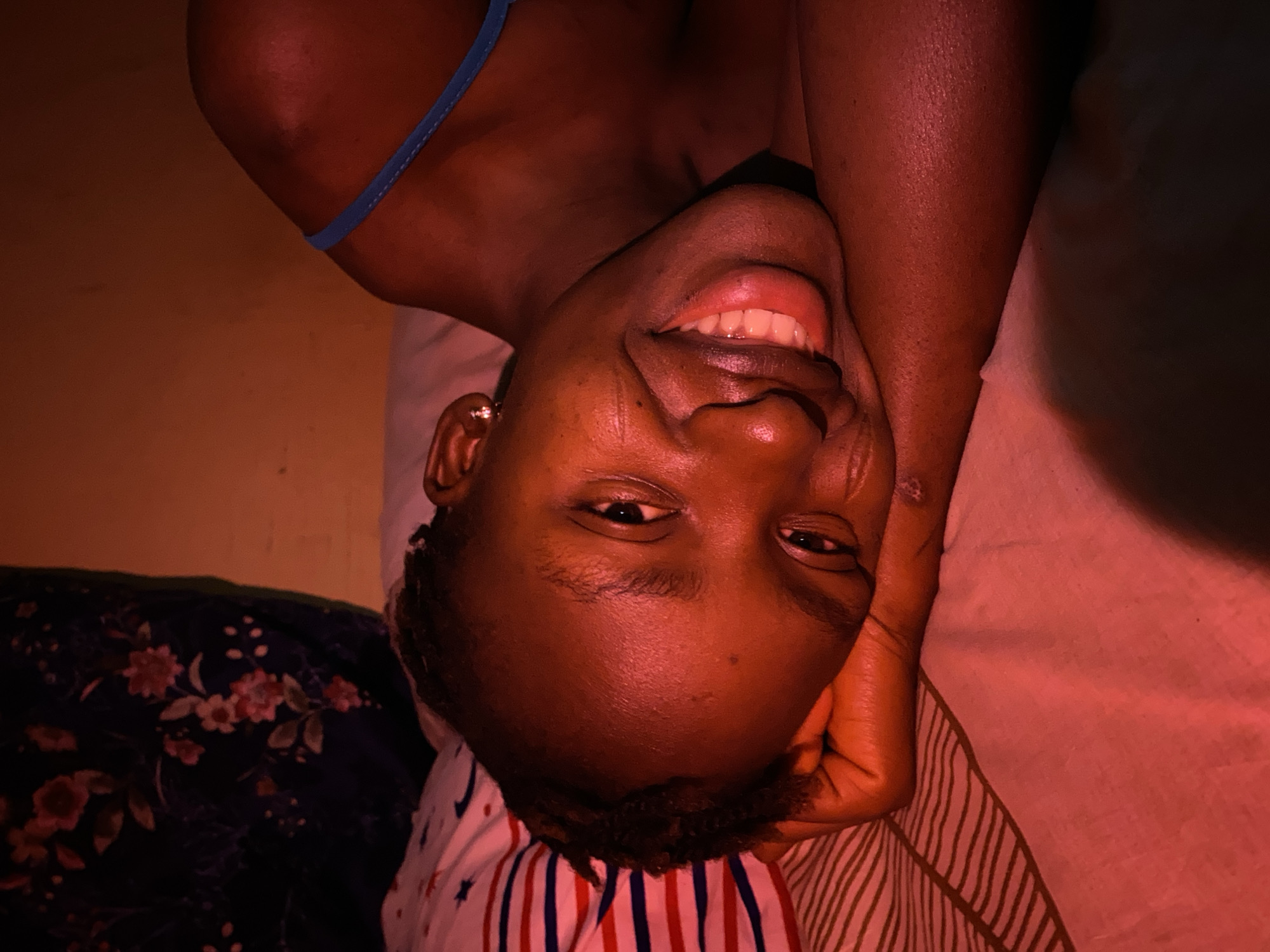 A young, dark-skinned woman lies on a bed with her head turned to the right and angled towards the camera. She is smiling, showing her teeth. Her right hand is propped up against her head. She is wearing a blue strap, possibly a part of her clothing. The bedding includes a pillow with red and white stripes and a blanket with a floral pattern. The background appears to be a wall with a warm tone. There is no way to know if this image was taken in Kuru Dungus, Nigeria or any other specific location from the given picture alone.