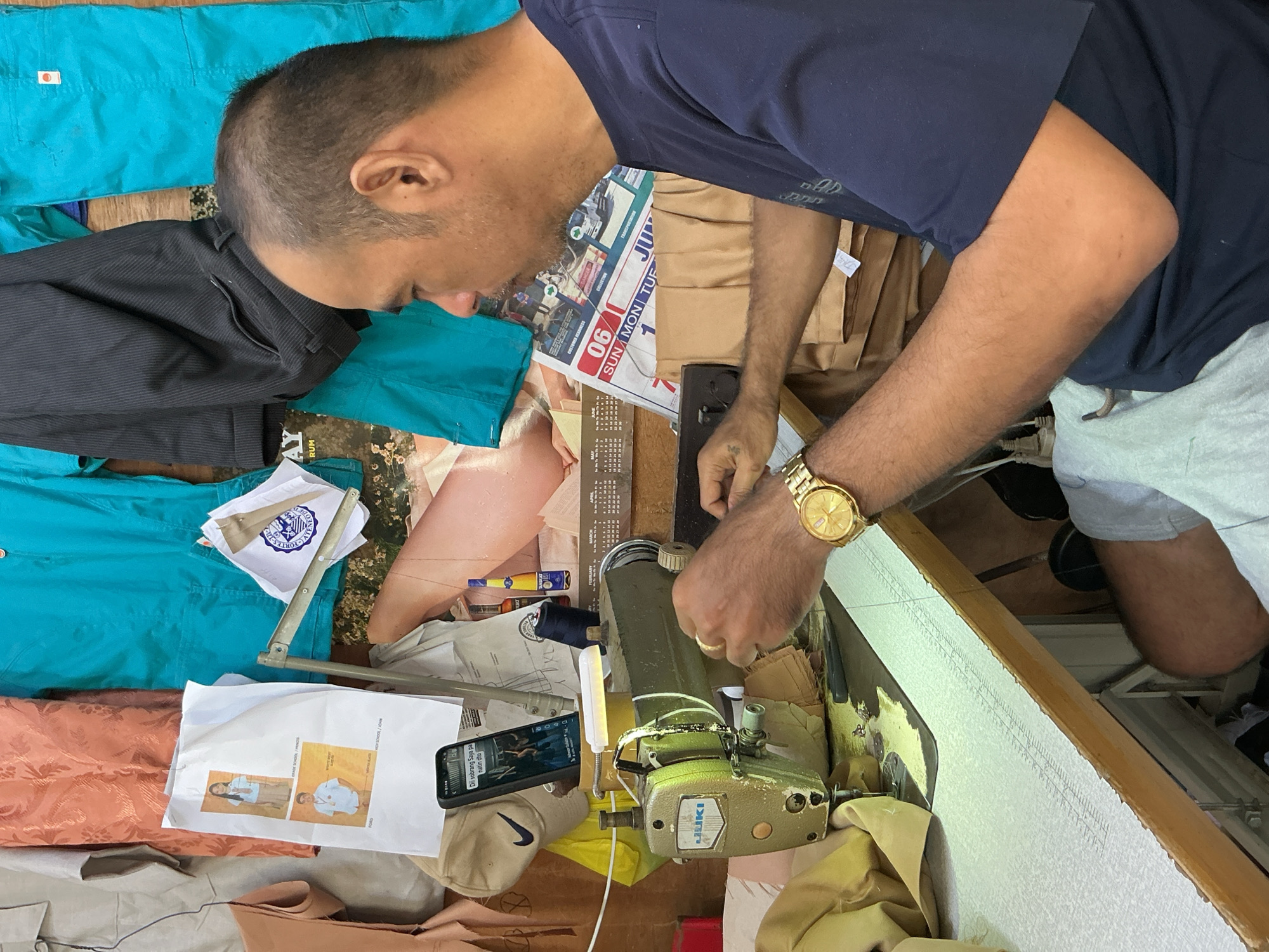 A middle-aged Filipino man is seen from the side, focused on operating a sewing machine in what appears to be a tailor shop. He is wearing a dark blue t-shirt and light-colored shorts, with a gold wristwatch on his left wrist. The sewing machine is a Juki model, green in color, positioned on a wooden table. The background is cluttered with various fabrics, garments, and other items, including a framed photograph of two young people, a phone displaying a video, and a calendar for June. The overall environment suggests a busy workspace. The image is set in Davao City, Philippines.