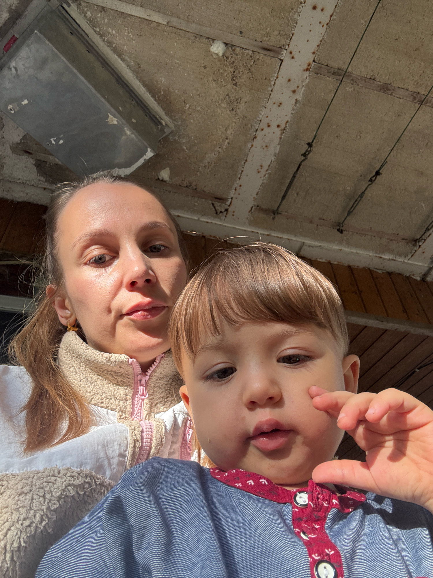 A woman and a young child are positioned in the foreground, facing the camera, with their upper bodies visible. The woman is on the left, her head tilted slightly. She has her hair pulled back in a ponytail and is wearing a light-colored jacket with a fluffy collar and pink zipper detail. The child is on the right, leaning into the frame, with light brown hair and is wearing a blue shirt with a patterned red collar. The child's right hand is raised towards their face. In the background, a rustic, unfinished ceiling structure is visible, with wooden planks and what appears to be an electrical conduit or fixture. The lighting suggests an outdoor setting, possibly under a covered porch or eaves. The image appears to have been taken in a residential area of Überherrn, Germany.