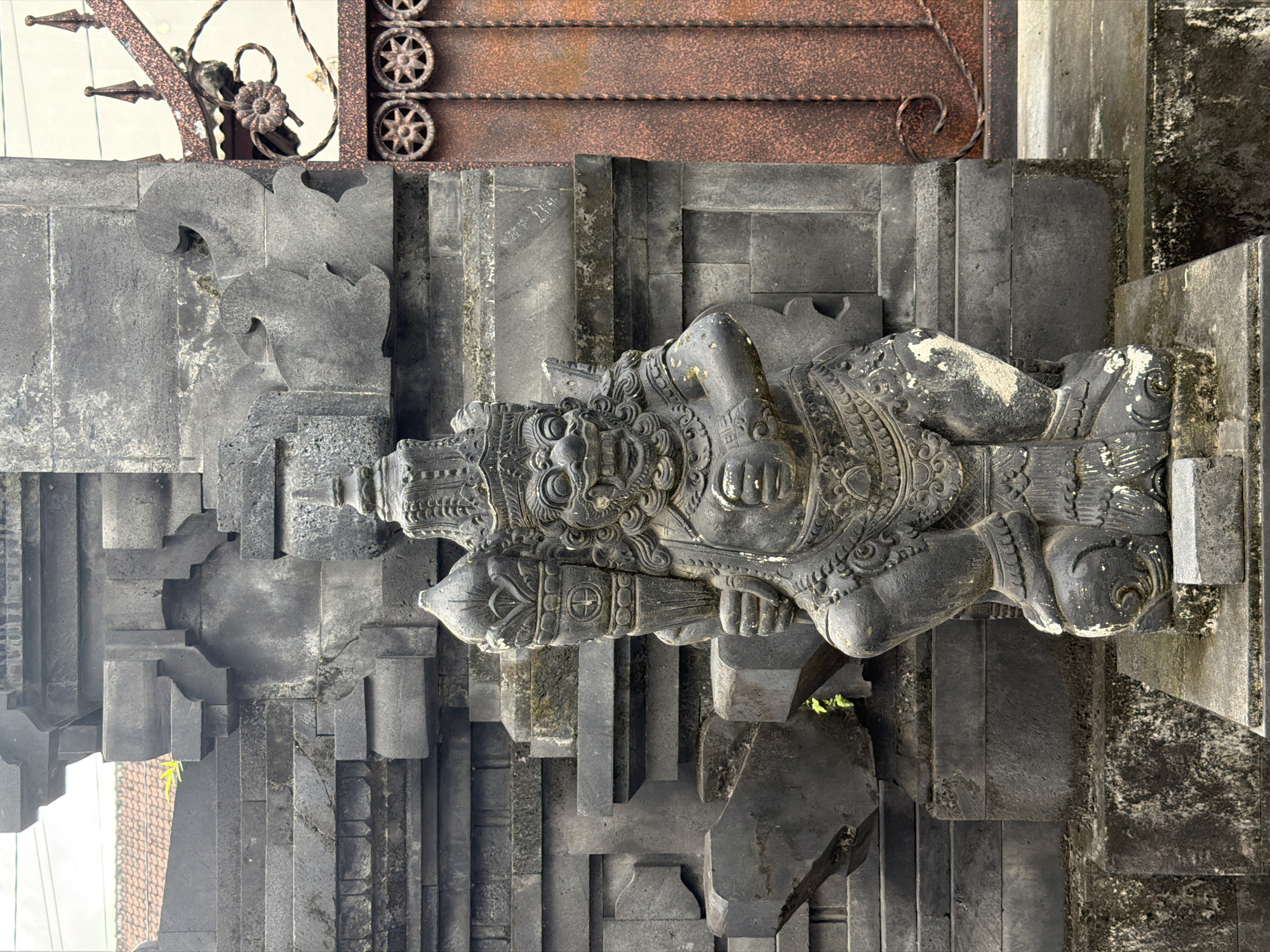 This image shows a stone statue in North Kuta, Indonesia. The statue is grey and depicts a mythical figure with intricate carvings and details. It is positioned on a stone structure composed of multiple steps and levels. Above the statue is a metal gate or decorative feature with a rusted or aged appearance. The surrounding environment appears to be an outdoor setting.