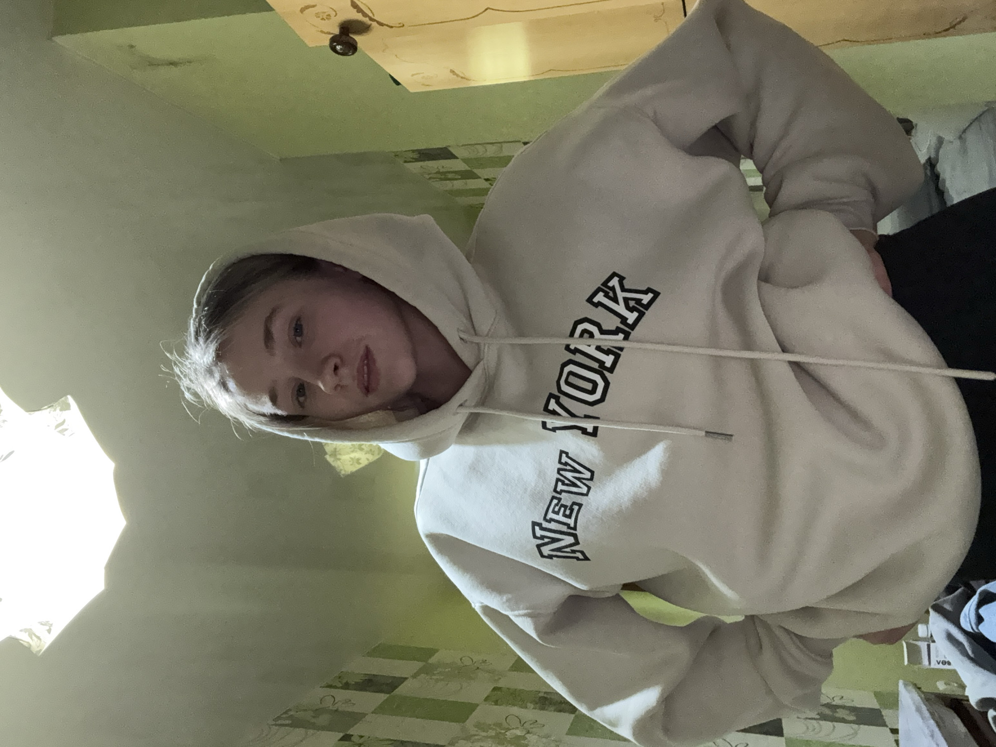 A young woman is shown in a hooded sweatshirt with "NEW YORK" printed on the front. She is positioned in a room with a green wall and tiled floor. The exact location in Izhevsk, Russia is not determinable from the image.
