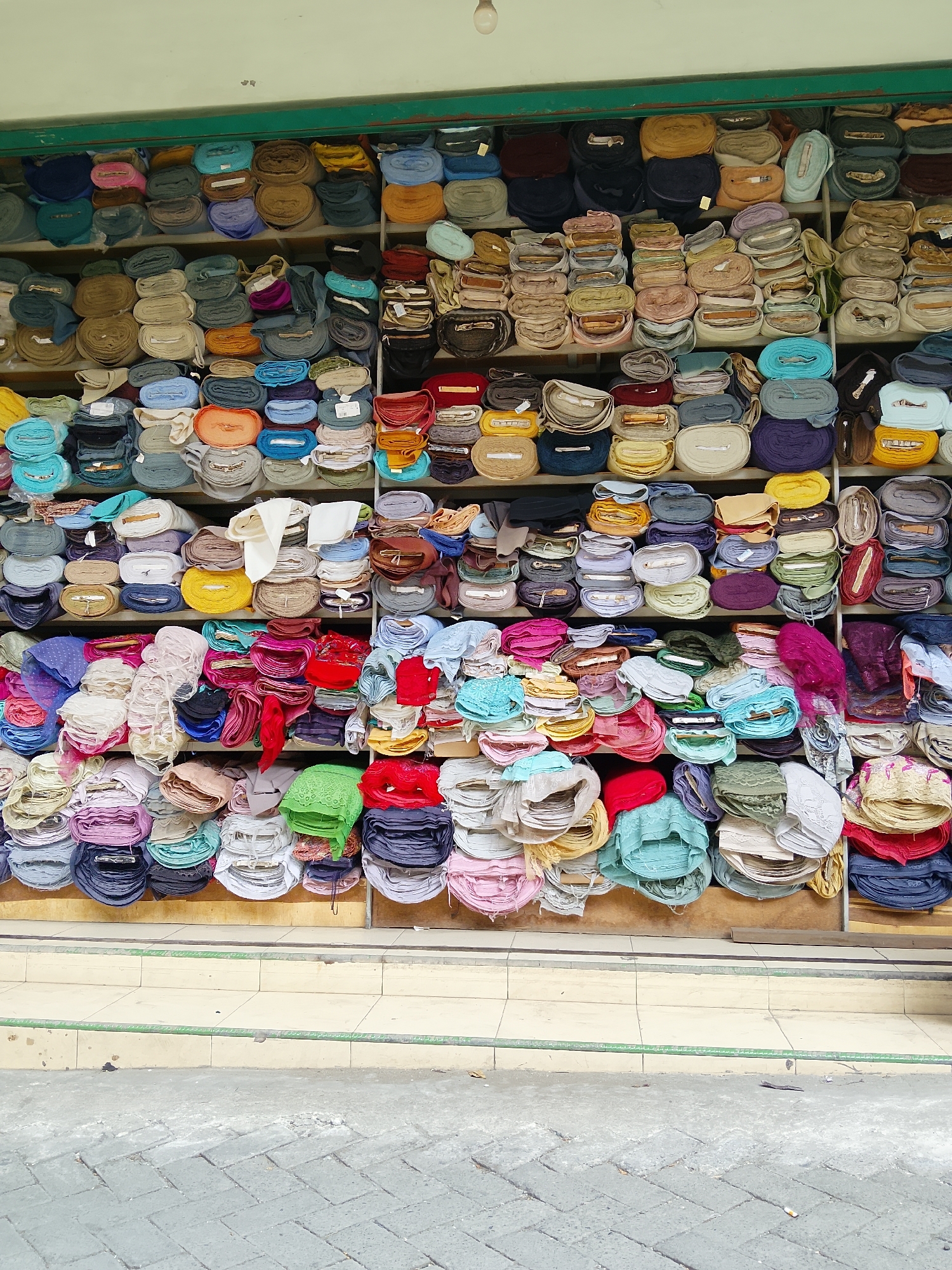 This is a head-on, eye-level shot of a stall displaying a large collection of rolled fabrics. The stall is located in Balun, Indonesia, as indicated by signage. The fabrics are neatly arranged on shelves, filling the frame from edge to edge. They come in a wide variety of colors and textures, including solid hues, patterned materials, and delicate lace. Some rolls are tied with string, while others appear to be loose. The background is a light-colored wall with a green trim at the top, suggesting an outdoor or semi-outdoor market setting. The foreground shows a tiled pathway, indicating the area in front of the stall. No people are visible in the image, and no explicit actions are taking place other than the static display of goods.