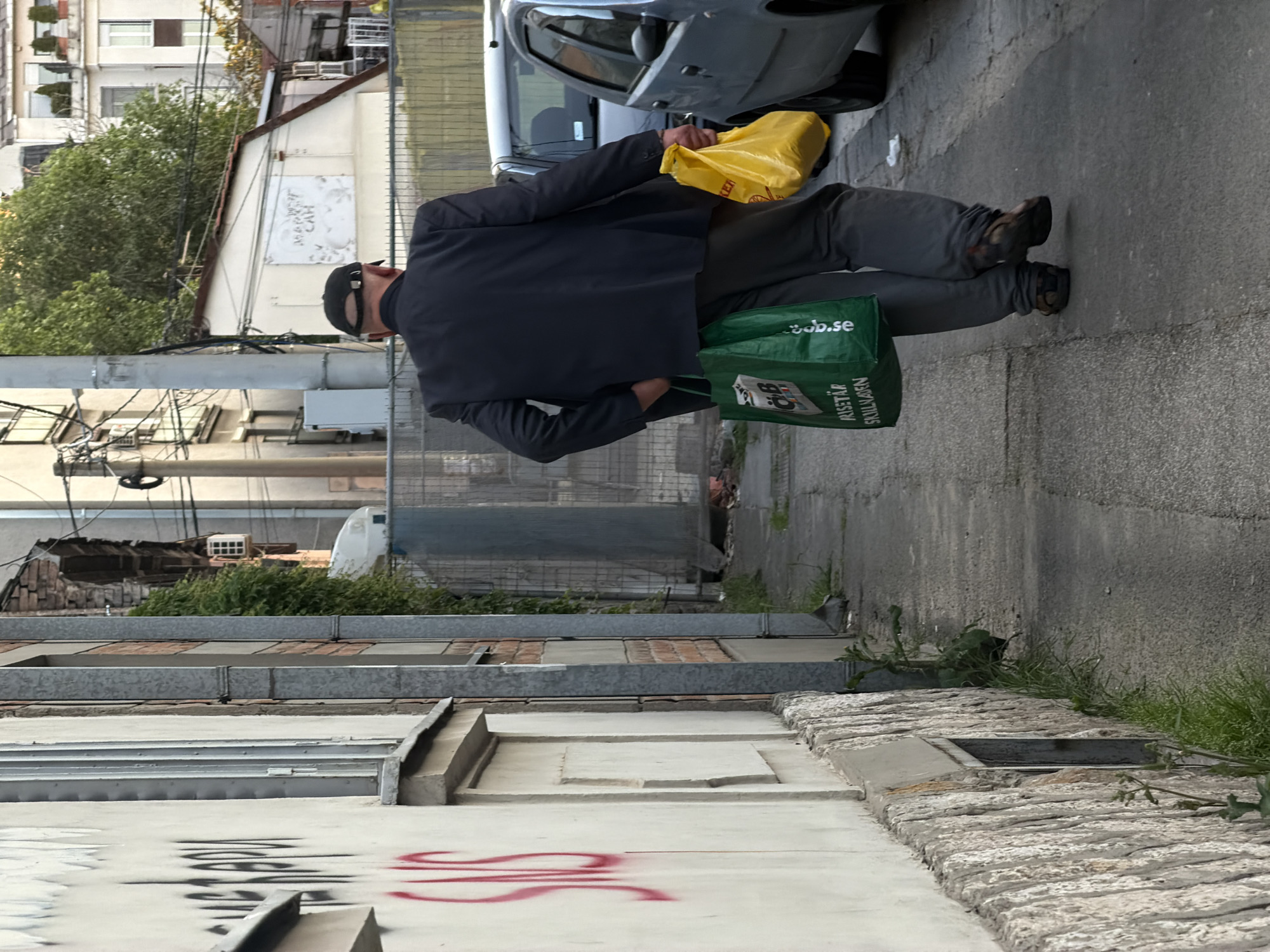 An image shows a man walking along a sidewalk with a yellow bag in one hand and a green bag in the other. He wears a dark suit jacket, gray pants, a black cap, and dark shoes. He is walking away from the viewer on a paved sidewalk adjacent to a street with a parked car. The sidewalk is flanked by a low wall and the side of a building with graffiti on it. Further in the background, there are buildings with greenery visible. The location is Vracar Urban Municipality, Serbia.