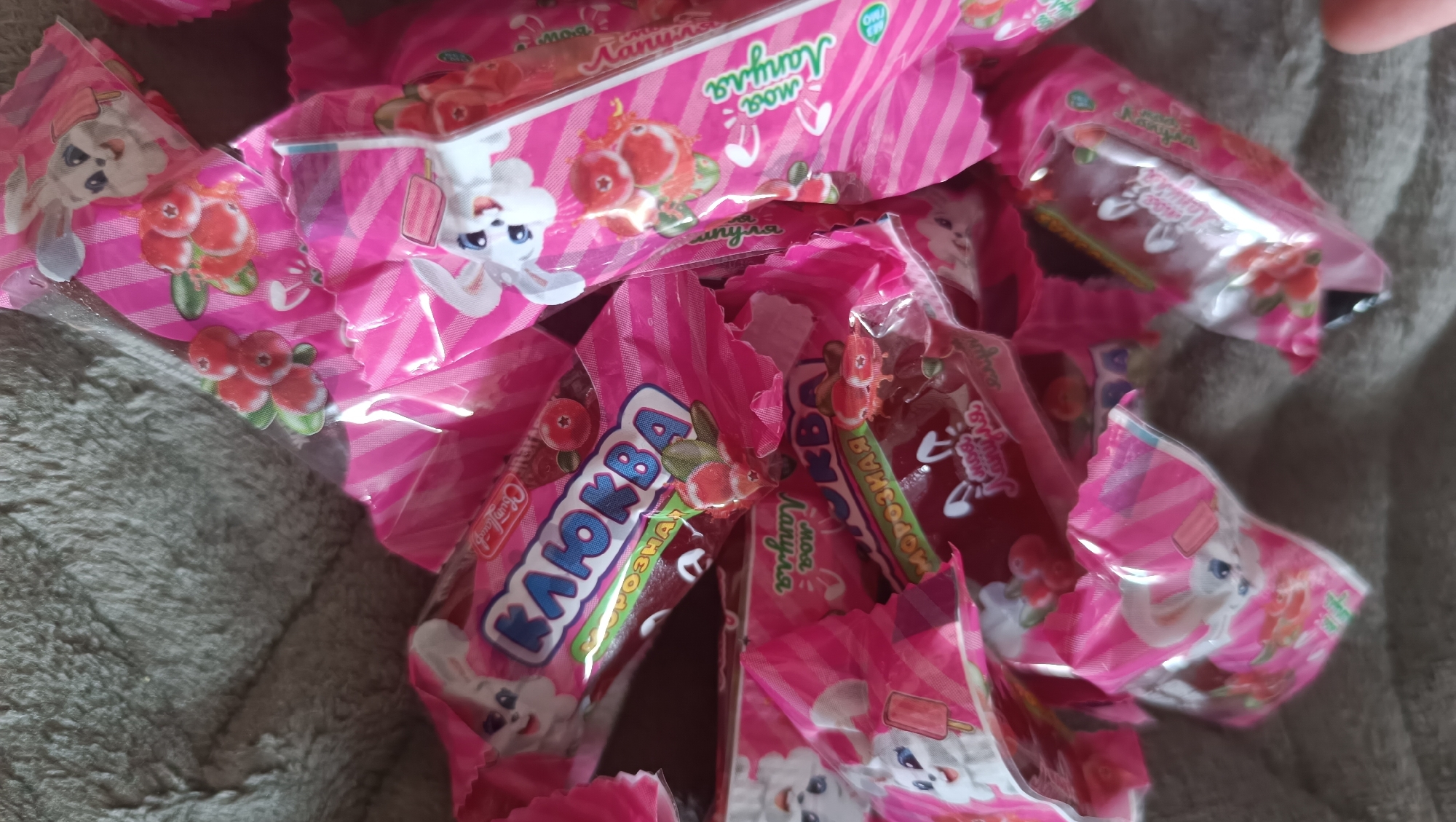 The image shows a pile of individually wrapped candies, predominantly pink with white striped packaging. The wrappers feature cartoon bunnies and images of berries, possibly cranberries, and some depict ice cream cones. Some of the text on the wrappers is in Cyrillic script. The candies are scattered on a textured, grey surface that appears to be fabric, possibly a blanket or towel. There are no discernible geographical markers or indications of the city of Karsha, Russia, in the image. The scene is static and focuses solely on the candy wrappers.