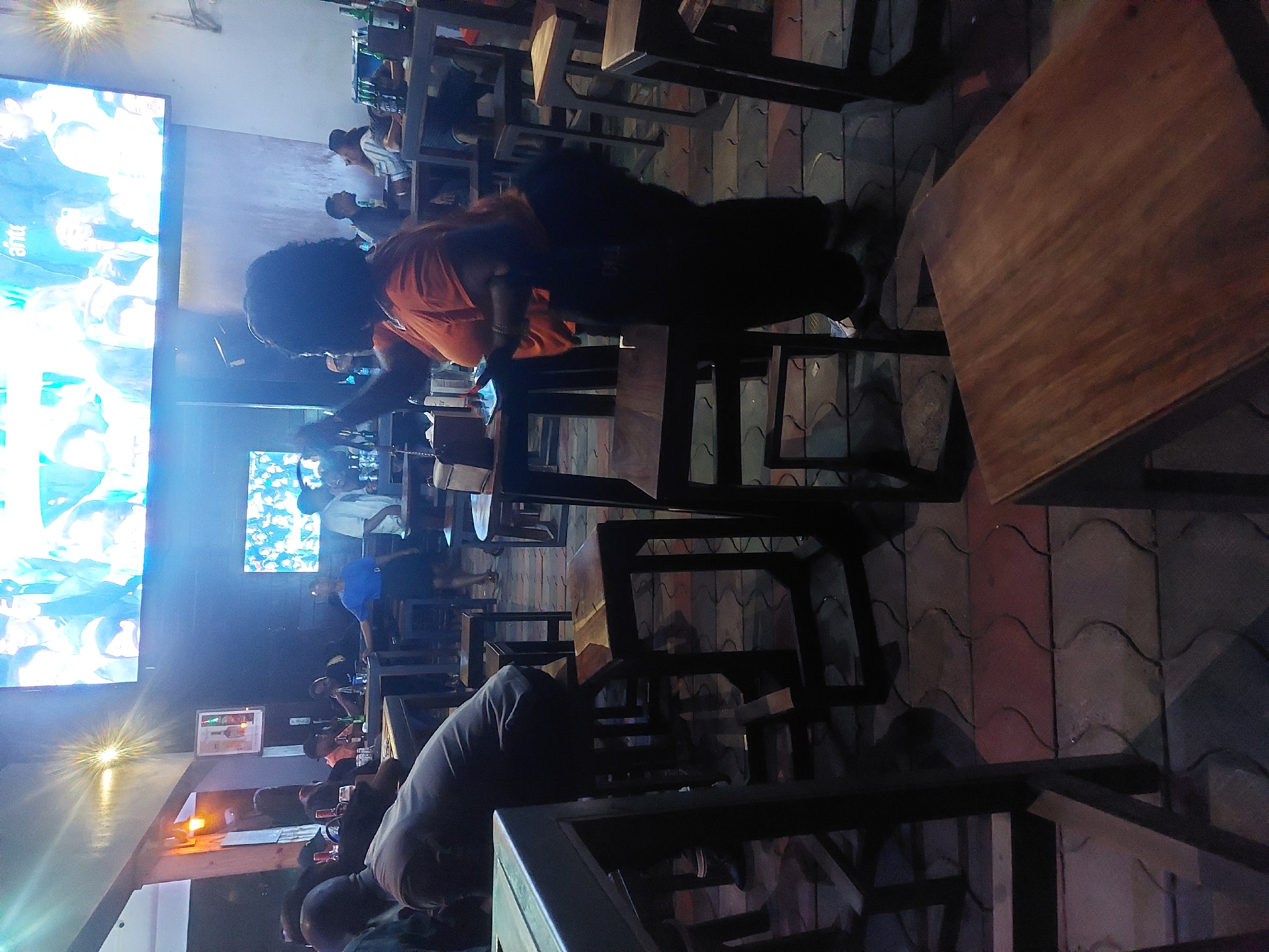 The scene depicts an active, dimly lit indoor or semi-outdoor bar or lounge establishment in the city of Dar es-Salaam, Tanzania, observed at night. The floor consists of interlocking concrete pavers in shades of red, grey, and dark tones. Two large display screens are mounted on a dark wall; the prominent upper screen shows televised entertainment or a sporting event featuring multiple figures, with the text "ahti" visible in its upper right quadrant.
The establishment is furnished with numerous dark wooden high-top tables and matching high stools. Approximately 10-15 individuals are present, predominantly adults. In the mid-ground, slightly to the right of center, a woman with dark, curly hair, wearing an orange t-shirt and dark pants, stands holding a smartphone in her left hand, appearing to be engaged with the device. A dark shoulder bag hangs on her right hip. To her immediate left, a man wearing a dark t-shirt is seated on a stool at a table, leaning forward with his back partially towards the viewer.
Further in the background, other patrons are seated or standing at additional tables; some tables display multiple beverage bottles, including green glass bottles. A woman in a blue top and dark skirt is visible standing near a table in the left background. Overhead artificial lighting and the luminous glow from the display screens provide illumination, creating a social gathering atmosphere.