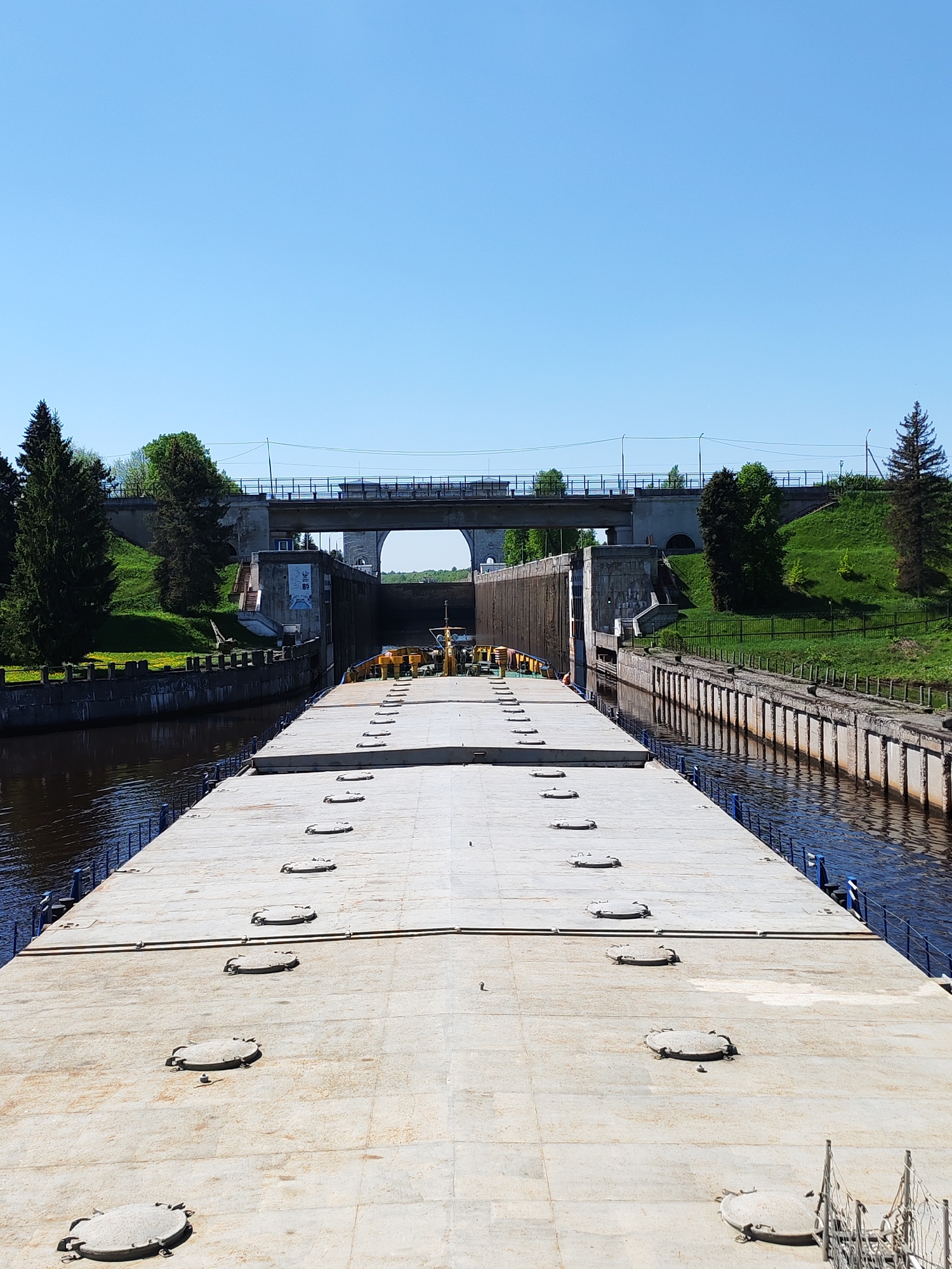 The image depicts a barge positioned within a canal lock system, situated in an unidentified location. The barge, characterized by a gray, flat surface punctuated by numerous circular hatches, dominates the foreground. The lock itself consists of concrete walls and structures. A bridge, with multiple arches, spans over the canal in the background. Verdant, grassy embankments line the canal banks, featuring trees and vegetation. The sky is clear and blue.