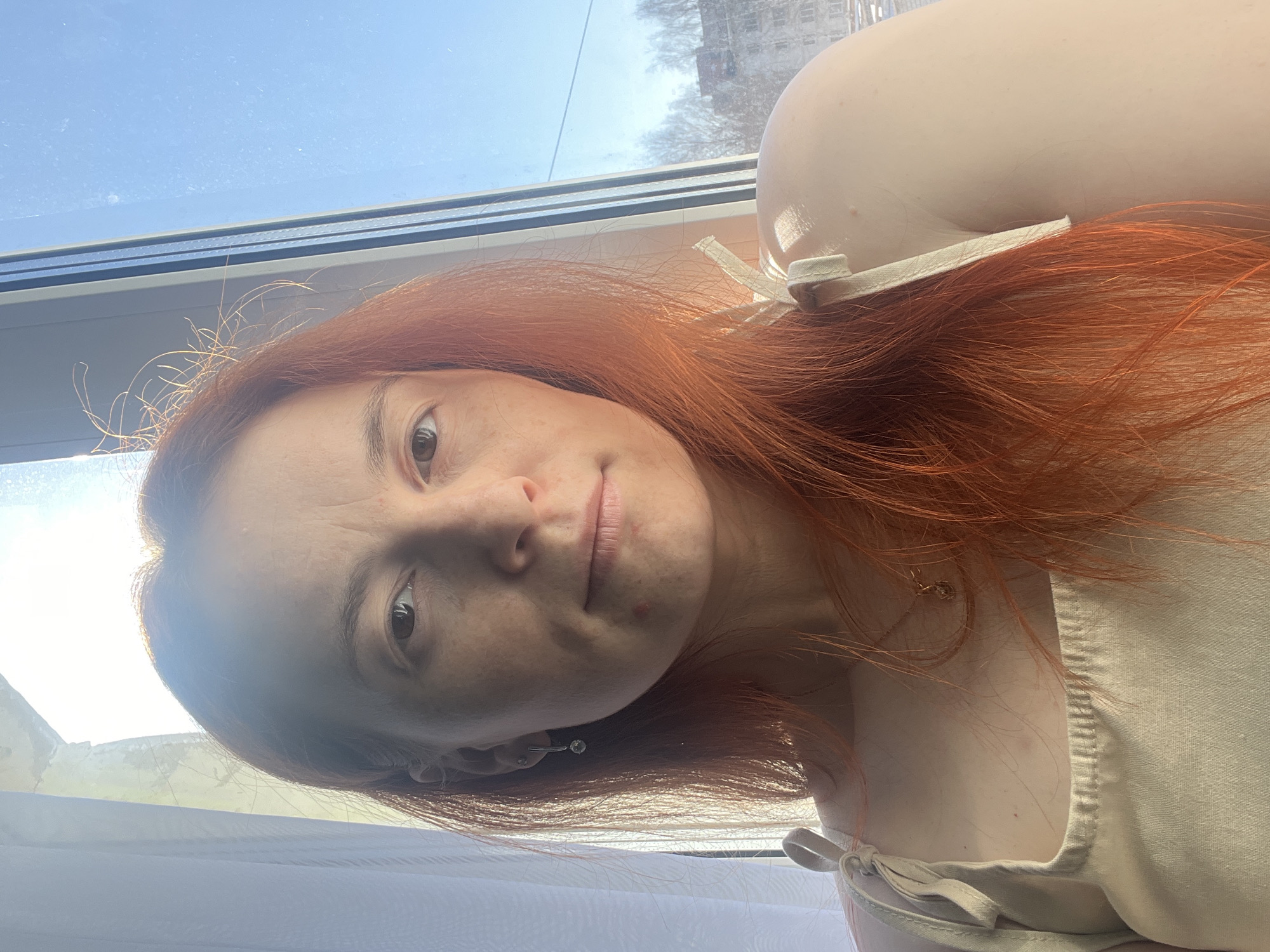A selfie taken indoors near a window. A woman with red hair is facing the camera. She has fair skin with freckles. She is wearing a light-colored top with ties at the shoulders. Her hair is slightly messy with flyaways. An earring is visible in her left ear. Outside the window, a building is visible, suggesting an urban setting. The city appears to be Chaikovsky, Russia, based on building characteristics and the presence of leafless trees, suggesting a possible late fall or early spring season. The window frame is visible, and the sky is blue.