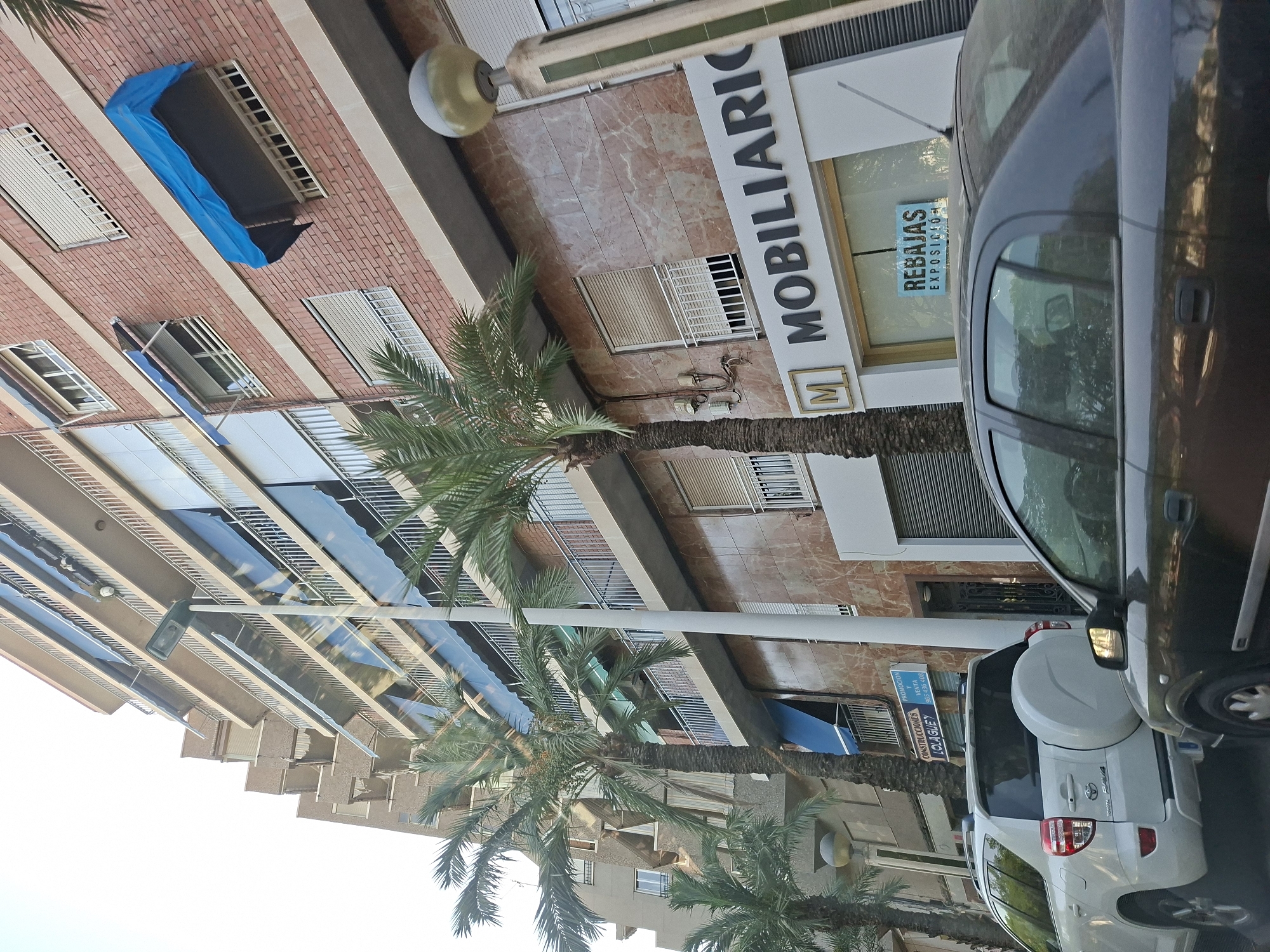 A low-angle, medium shot captures a street scene in Elche, Spain, featuring a multi-story brick building with balconies and awnings, palm trees, and cars. The building's facade is a mix of brick and marble-patterned tiles. Several balconies on the upper floors are adorned with blue retractable awnings. A street lamp stands on the right side of the image. In the foreground, two cars are visible: a white SUV with a spare tire mounted on the rear, and a dark-colored sedan partially obscuring the SUV. A white sign with the text "MOBILIARIO" is mounted on the lower section of the building. Smaller blue signs with Spanish text, "CONSTRUCCIONES LOLA GUEY" and "PROMOCION VENTA", are visible on the left side of the building. Another blue sign with "REBAJAS EXPOSICION" is placed on the roof of the dark-colored sedan. The overall scene depicts a commercial area with residential elements.