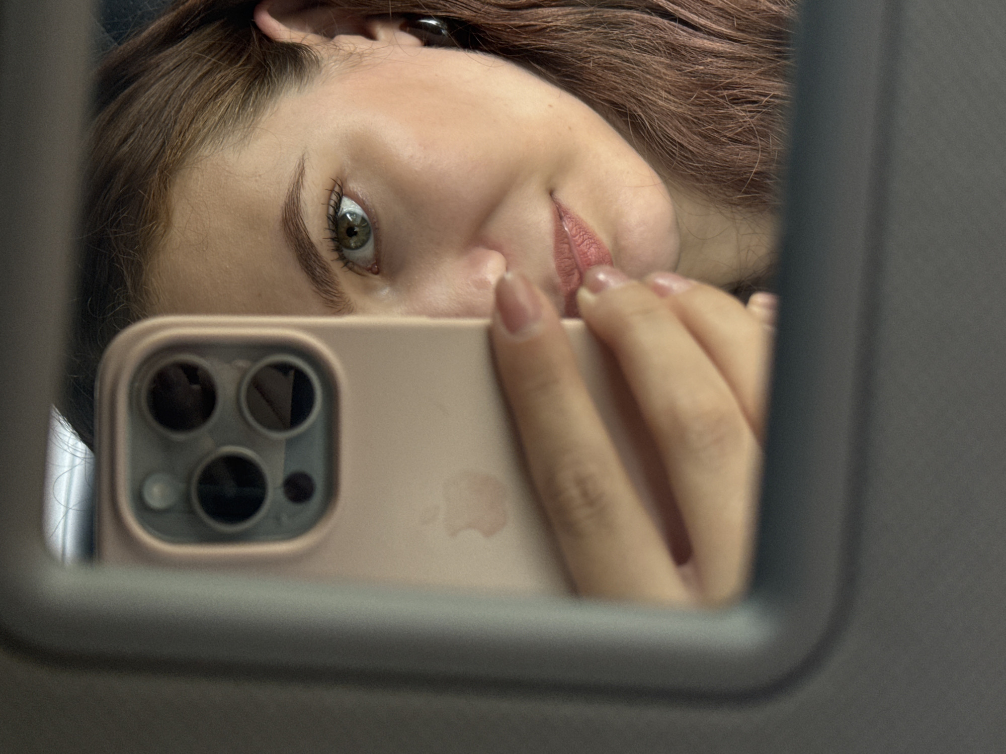 A young woman is reflected in a car's visor mirror, holding a smartphone to take a selfie. She has light brown hair, green eyes, and is wearing makeup. Her left hand is visible, holding the phone. The phone is a light pink iPhone with a triple camera system. The mirror frame is dark gray and appears to be part of a car interior. The background is blurred. No specific location in Chaikovsky, Russia is identifiable.
