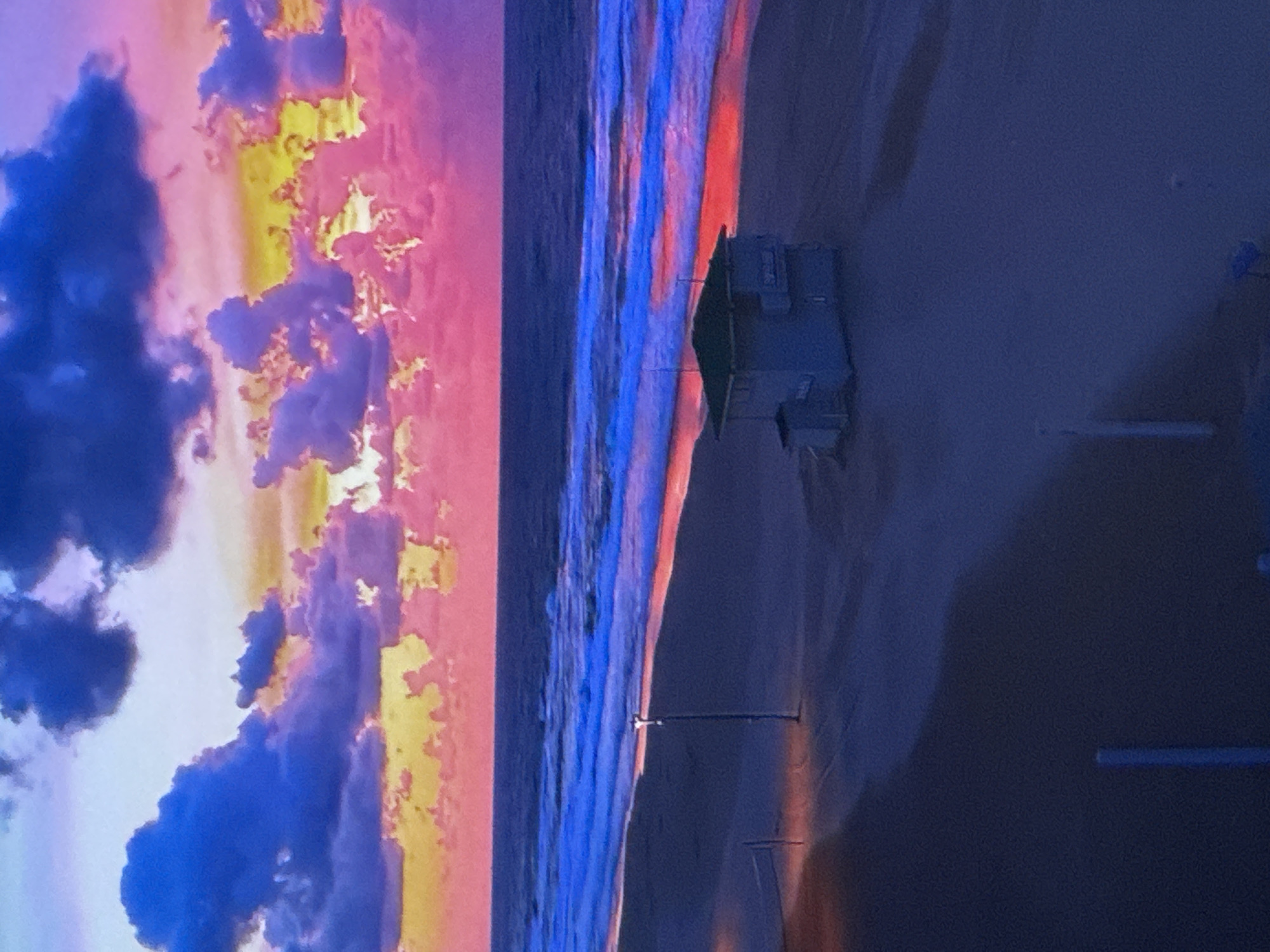 A beach scene at sunset features a vibrant sky with streaks of pink, yellow, and deep blue clouds. The ocean is a dark blue with bright blue and red-tinged waves breaking on the shore. On the sandy beach, a small, light blue lifeguard tower with a green roof stands near the water's edge. Further back on the beach, a single lamppost illuminates the sand. A volleyball net is visible in the distance. The overall impression is a serene, yet dramatically lit coastal environment. The location is the city of Unknown, Russia.