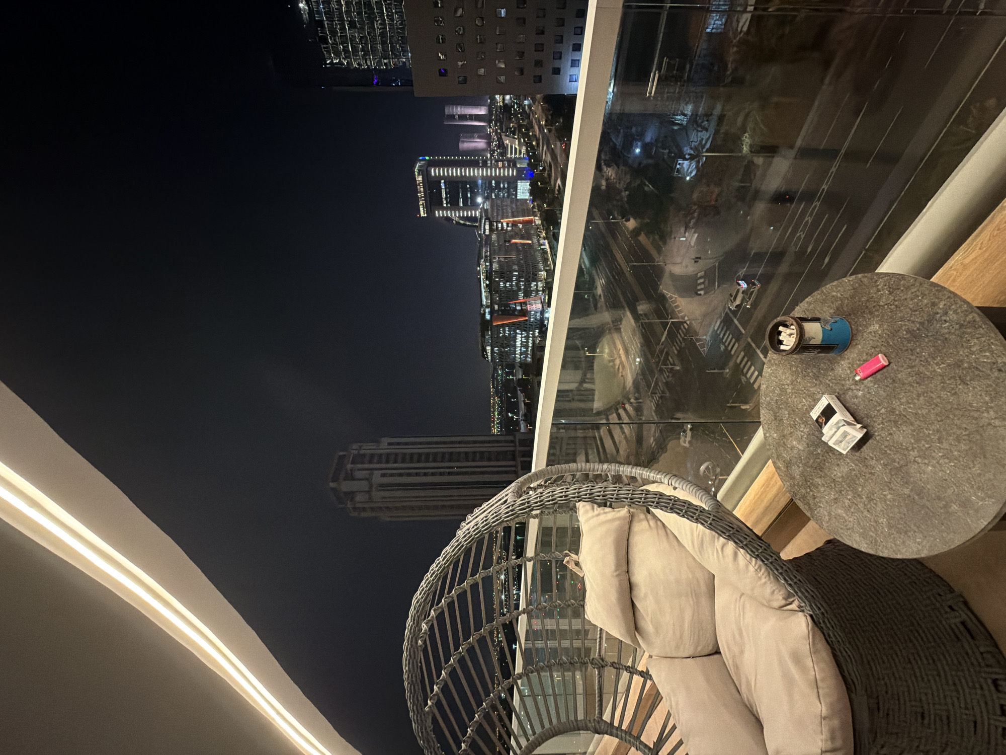 An outdoor patio is shown at night, overlooking a city skyline. In the foreground, a round, grey, textured table holds a small pack of cigarettes, a lighter, and an ashtray with cigarette butts. Next to the table is a large, woven armchair with light-colored cushions and a dark grey throw blanket. A white railing with a glass barrier separates the patio from the view of the city.
The city skyline features numerous illuminated skyscrapers and buildings, indicating a bustling urban environment. A multi-lane road is visible below the balcony, with streetlights and moving vehicles. The sky is dark, suggesting it is nighttime. The overall mood is peaceful and relaxed, with the ambiance of a city at night. No people are visible in the frame. There is no visible text in the image.