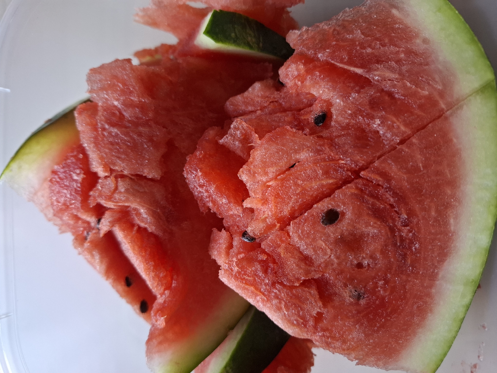 A close-up, high-angle shot features several slices of ripe watermelon, some with seeds visible. The watermelon flesh is a vibrant red, with a lighter pinkish-white rind. The texture of the watermelon is somewhat rough and juicy. The image is set against a plain, light-colored background. There are no people or any recognizable landmarks indicating a location in Tyumen, Russia.