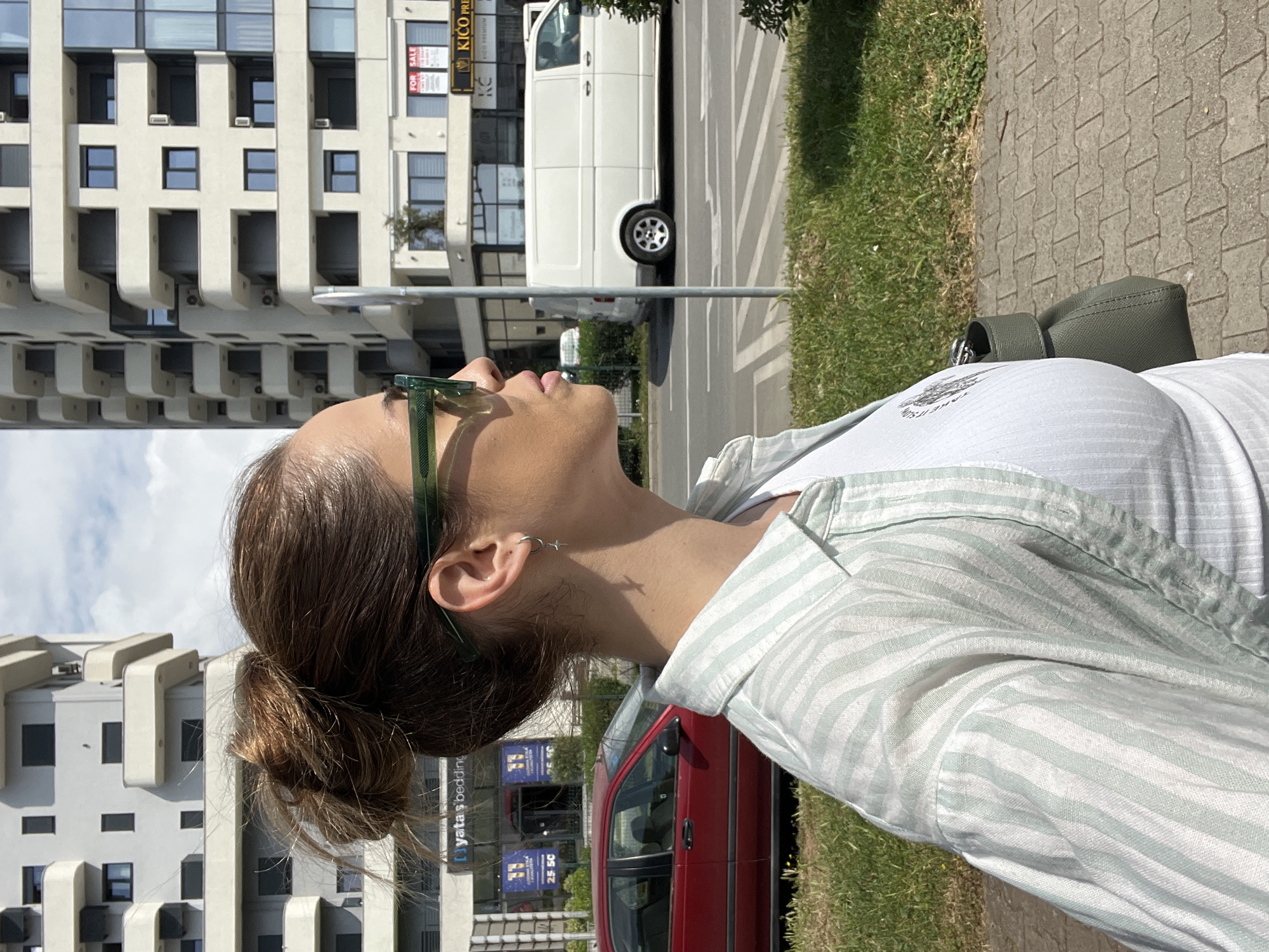 An outdoor, brightly lit image, oriented on its side, shows a person from the chest up in the foreground, with an urban setting behind them. The person has brown hair styled in a bun, wears green-tinted sunglasses on their head, a white t-shirt with partial text visible, and a light green and white striped button-up shirt over it. Their head is tilted back, looking upwards, revealing the right side of their face and neck. Multiple small piercings are visible on their right ear, including a hoop and a cross-shaped dangle.
The background features several multi-story buildings with modern architectural designs, including distinctive protruding balconies or facade elements. A street with a white commercial van and a red sedan is visible between the foreground subject and the buildings. A paved sidewalk and grassy areas run along the street. The bright sunlight suggests it is daytime, casting clear shadows.
Visible text includes "KICO PREMIUM" and "FOR SALE" on a sign attached to one of the buildings. Another sign on a different building reads "yata's bedding". On the person's white t-shirt, the words "TAKE IT" are partially visible.