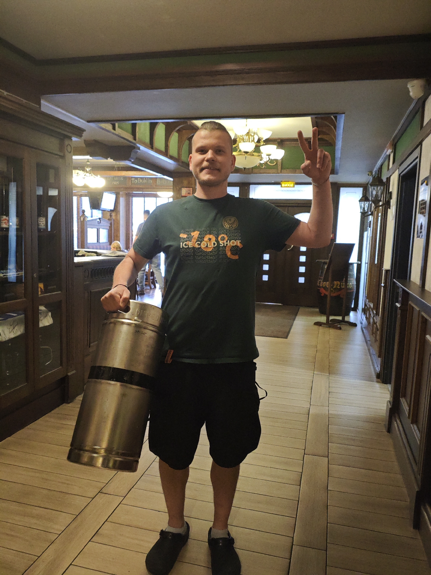 A man stands in a hallway that appears to be inside a bar or restaurant. He is holding a silver keg in his left arm and is making a peace sign with his right hand. He is wearing a dark green t-shirt with orange lettering that reads "ICE COLD SHOT 789" and black shorts. The hallway has wooden paneling on the right side and wooden cabinets on the left. There are lights and decorative elements on the ceiling. The flooring is made of light-colored tiles laid in a diagonal pattern. In the background, there are doorways and other patrons visible. The man's hair is styled in a short buzz cut with the top section slicked back. He has a mustache and a faint smile. There is no indication of the city of Kyiv, Ukraine.