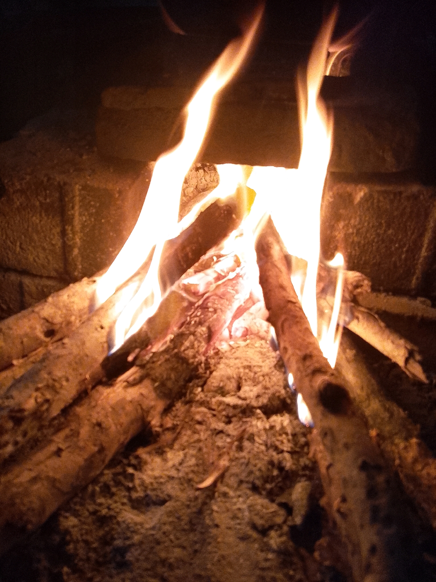The image depicts a close-up view of a campfire. Several logs are arranged in a fire pit, with flames engulfing parts of the wood. The background appears to be a brick structure, possibly part of a fireplace or hearth. The lighting is dim and warm, emanating from the fire itself, creating strong highlights and shadows. There is no indication of the city of Wadang, Indonesia, or any people present in the scene.