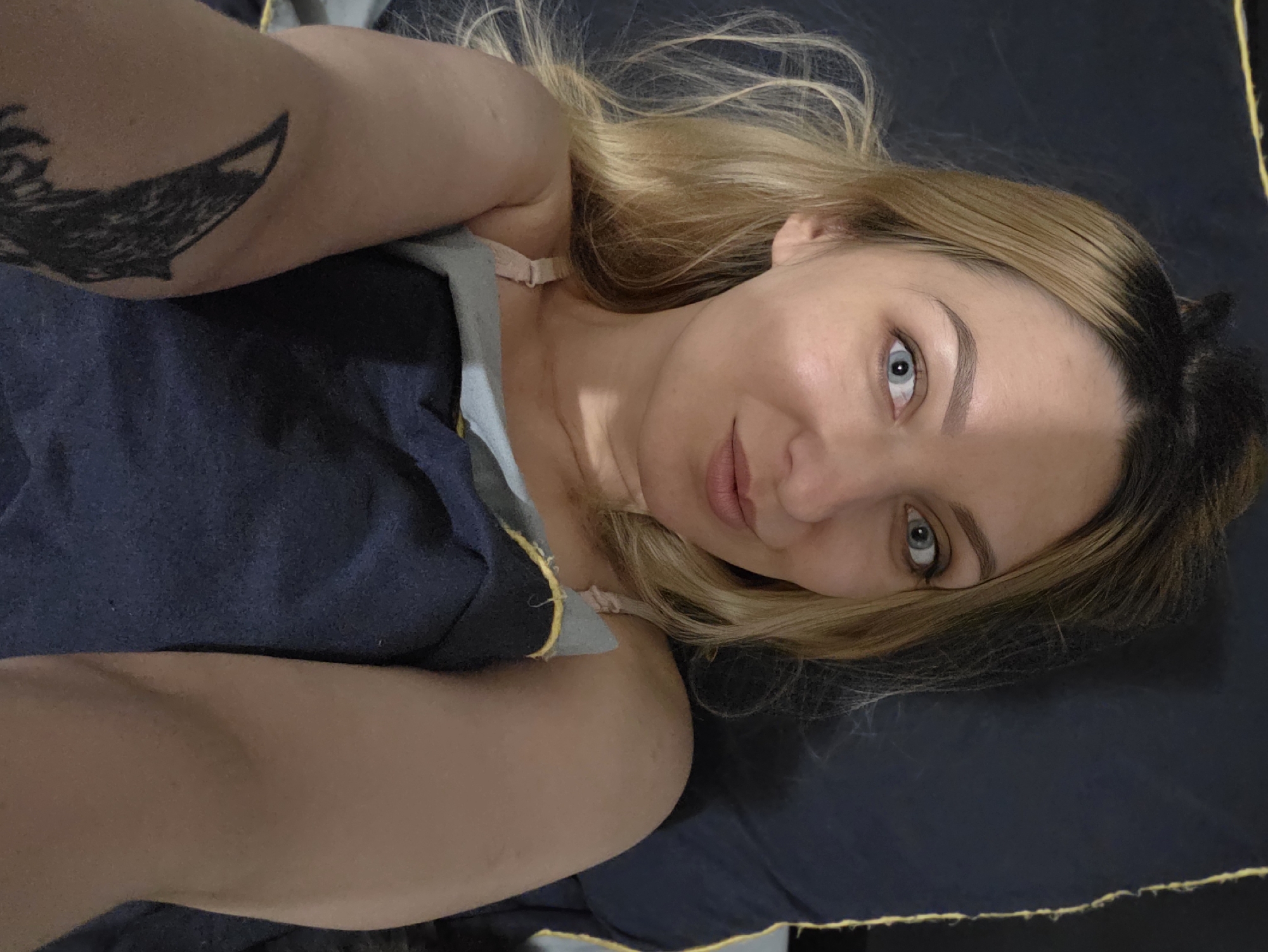 A blonde female is depicted in a selfie-style shot, lying on her back on a dark blue pillow. Her blue eyes are wide open, gazing directly at the camera, with a slight smirk on her lips. Her blonde hair with darker roots is loosely pulled back, and some strands fall around her face. A light-colored bra strap is visible on her left shoulder. Her body is partially covered by a light blue-grey sheet, beneath a dark blue blanket with visible yellow stitching or trim along its edge. A black, stylized tattoo is visible on her right upper arm. The background is a solid dark blue, matching the bedding, with a yellow vertical line on the right side of the frame. The lighting creates a shadow across the left side of her face. The image provides no visual information to confirm a location in Minsk, Belarus.
