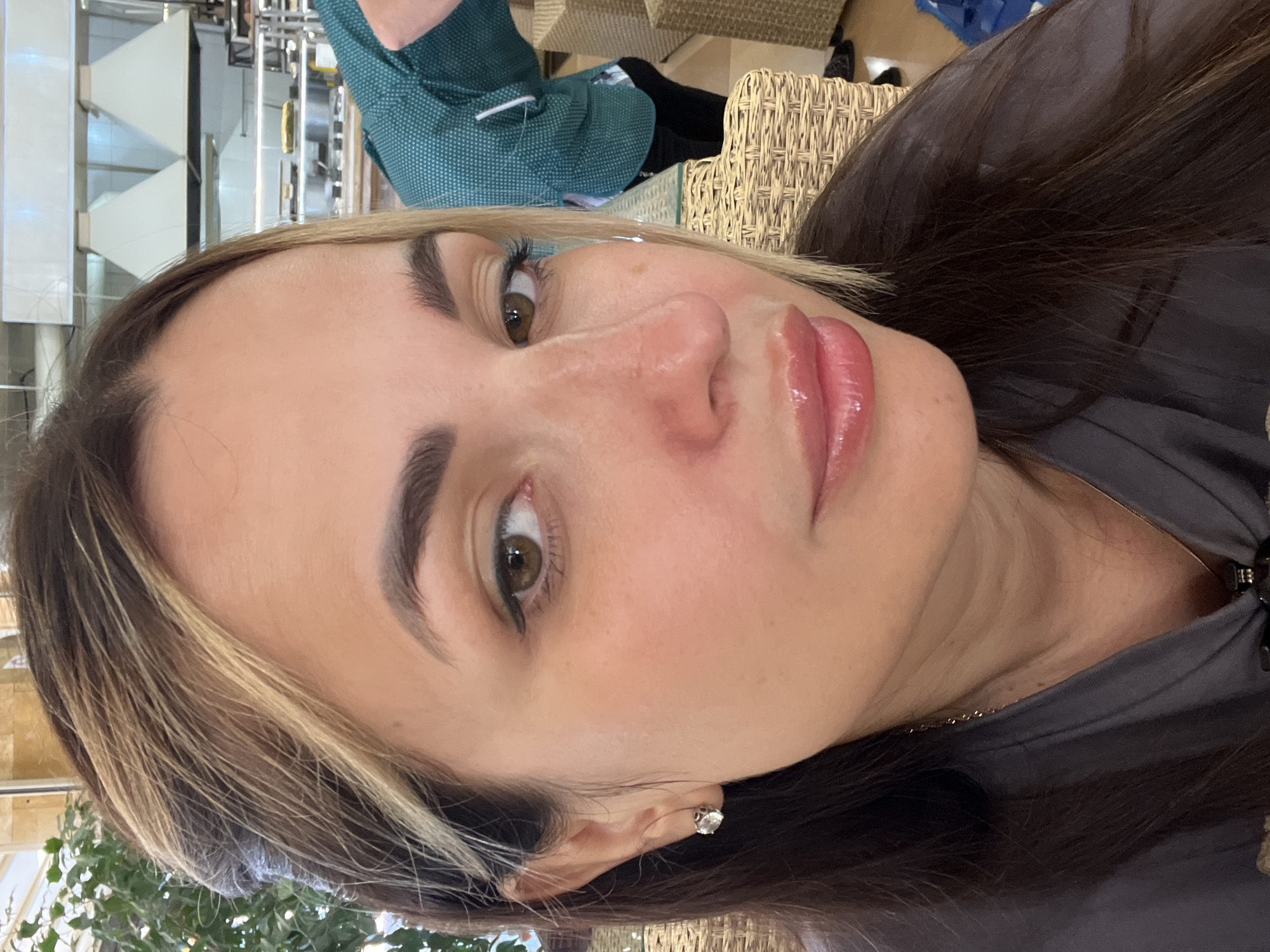 A medium shot from the chest up shows a woman with dark brown hair with blonde highlights, dark eyebrows, and brown eyes. She is wearing a grey top, a gold necklace, and a stud earring in her left ear. Her lips are glossy pink. She is looking towards the camera. In the background, a person in a green shirt is visible, and restaurant equipment is present. The setting appears to be a restaurant or cafe with wicker chairs.