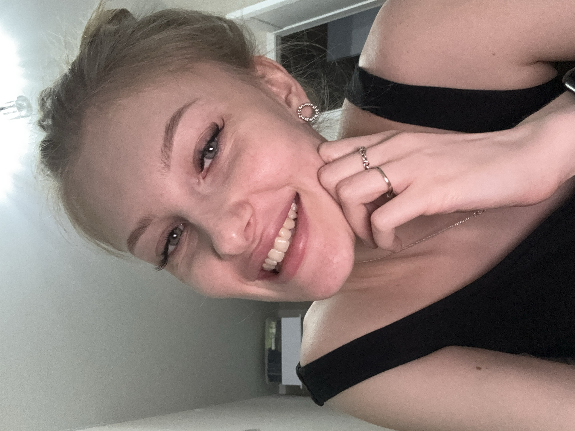 A smiling young woman with blonde hair pulled back is the primary subject of this portrait-style image. She is wearing a black, off-the-shoulder top and several rings on her left hand, which is resting against her cheek. She is also wearing circular earrings and a delicate necklace. The lighting is bright and appears to be artificial, possibly from an overhead light.
The background is minimal and appears to be an interior space, perhaps a room in a house or apartment, with neutral colored walls. Through an open doorway in the background, a glimpse of another room is visible. The image location cannot be determined based on available visual information alone, and associating it with the city of Perm, Russia, is not possible without additional data.