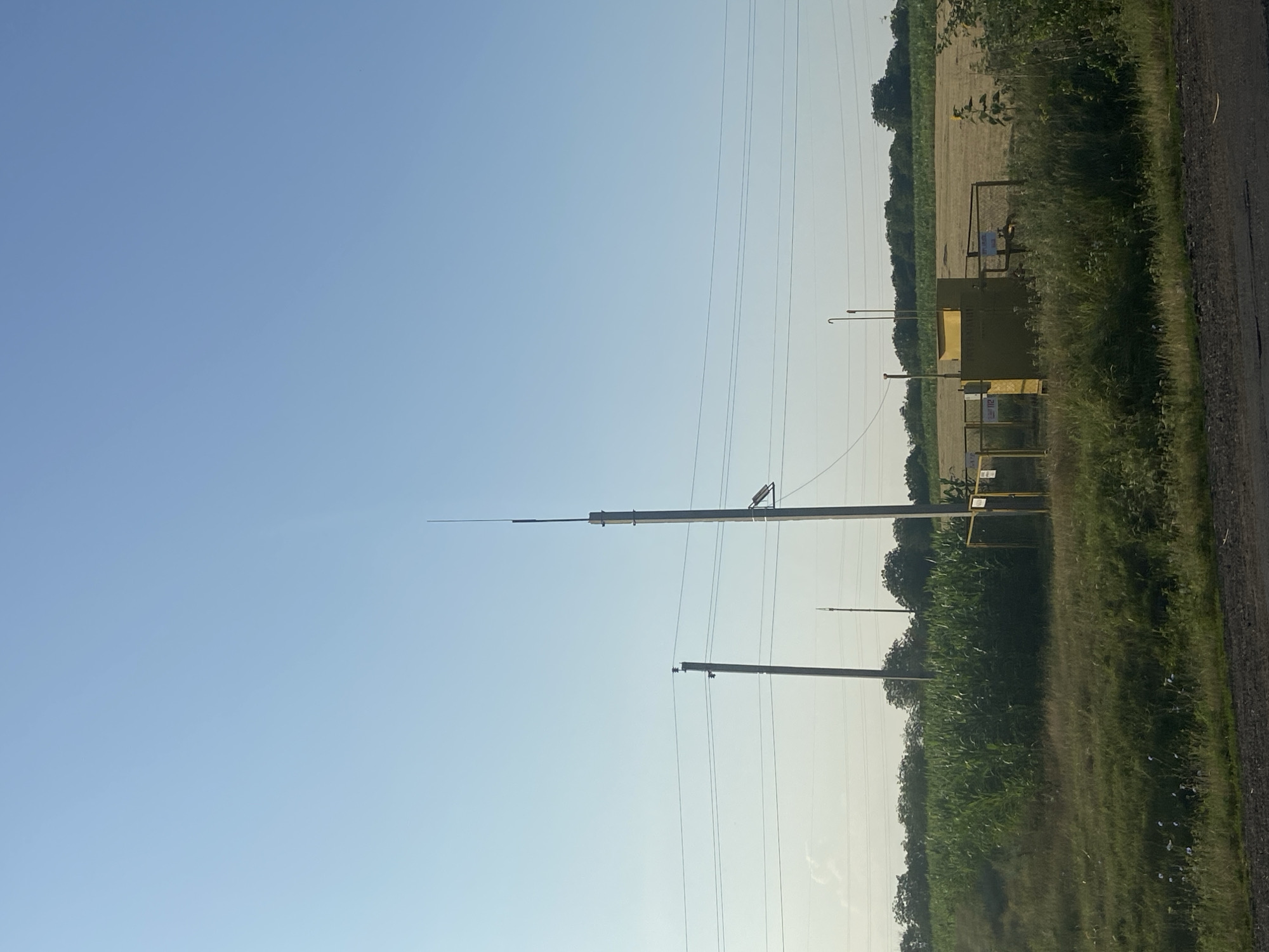 An outdoor scene depicts a clear sky over a rural landscape with agricultural fields. In the foreground, a paved road runs horizontally. To the left, tall green corn stalks are visible. To the right of the cornfield, a yellow industrial enclosure, possibly a gas regulator station, is situated behind a chain-link fence. The enclosure has two vertical pipes extending upwards and a warning sign on the fence. A tall utility pole with a lightning rod stands behind the enclosure. Further in the background, a line of trees separates the fields. Another utility pole is visible to the left of the tall one. The overall impression is one of a utility infrastructure element within an agricultural setting.