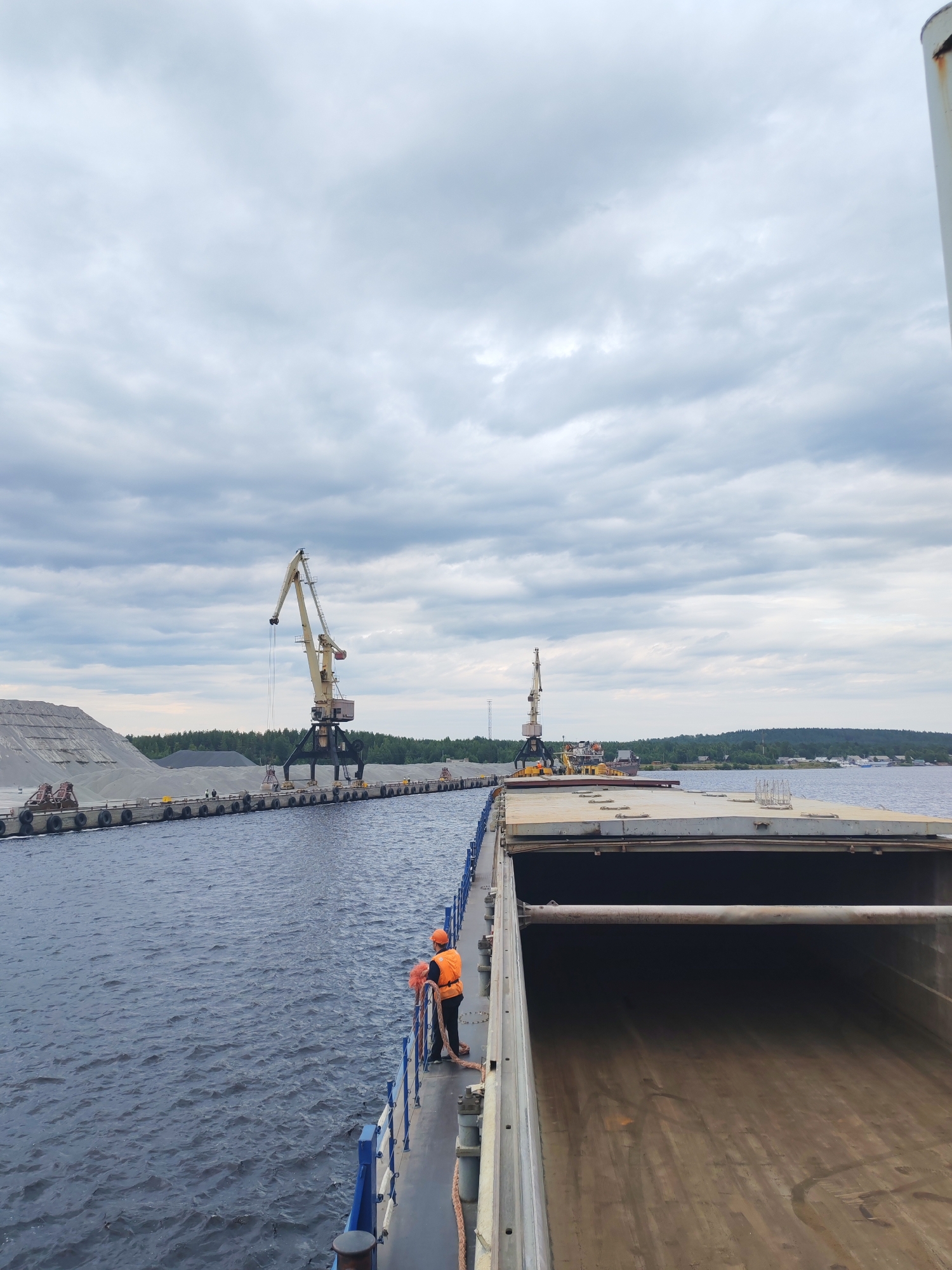 A person in an orange vest and hard hat stands on the deck of a cargo ship in Shalsky, Russia, looking out over a body of water. The ship is at a dock where large piles of gravel and two industrial cranes are visible. The sky is overcast with gray clouds. The water is dark and choppy, and the surrounding area is forested. The scene appears to be a working port or industrial site.