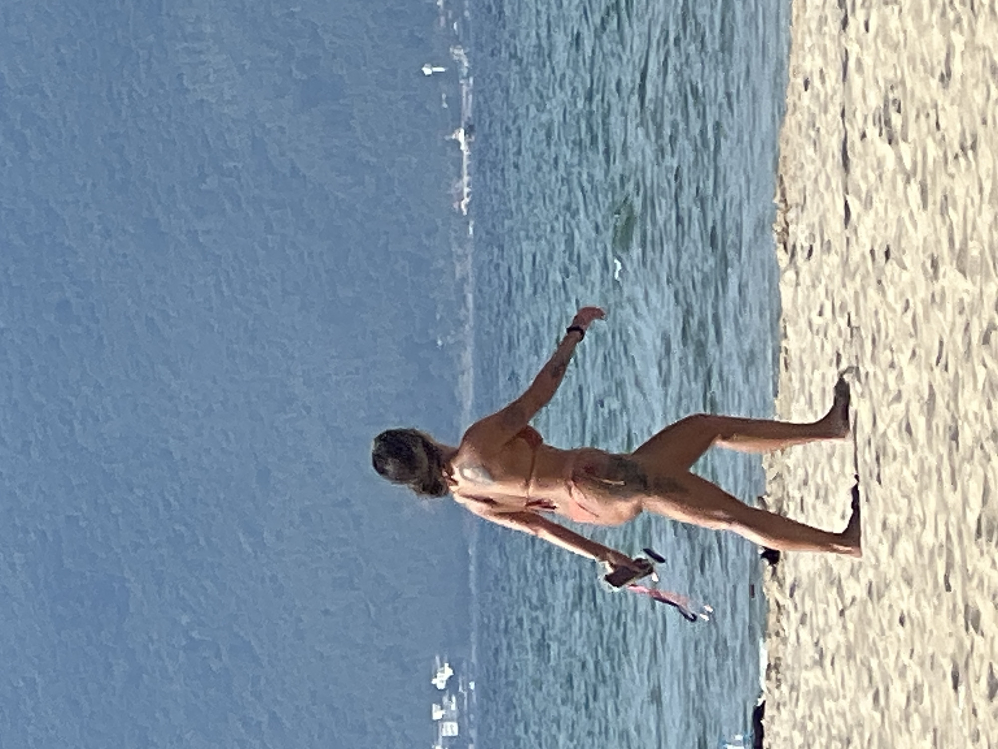 A lone woman walks along a sandy beach with the ocean to her right and a densely forested, mountainous coastline in the distance. She is wearing a rust-colored bikini and walking away from the camera towards the right of the frame. In her right hand, she is holding a pink and black object, possibly a pair of flip-flops or sandals. The woman has a tattoo visible on her left thigh and a sun tan. The water is a vibrant blue with small waves lapping the shore, and the sky is a clear, pale blue. Distant buildings are visible on the coastline in the far left of the image. This scene is consistent with the beaches of Đà Nẵng, Vietnam, known for their sandy shores and scenic mountain backdrops.