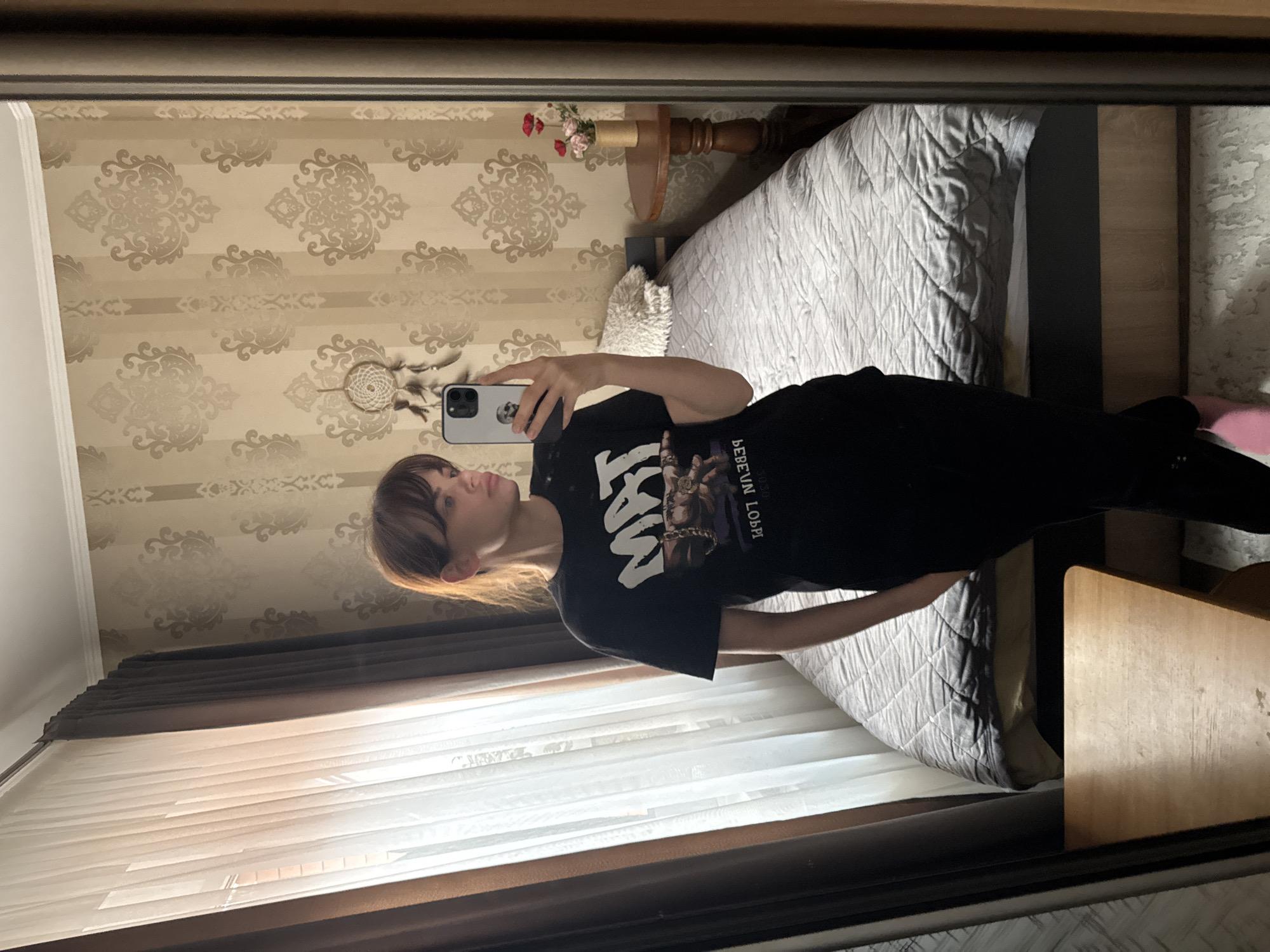 The image shows a reflection of a young person, likely female, taking a mirror selfie in a bedroom in Rostov-on-Don, Russia. The person has light brown hair with bangs and is looking into the camera of their smartphone, which they hold up with their right hand. They are wearing a black short-sleeved t-shirt with a graphic print featuring a figure wearing a chain. The visible text on the t-shirt, when unreversed from the mirror image, reads "БЕБЕМ ЛОРГ" (BEBEM LORG) and "MMX". They also wear dark trousers, and a pink slipper or sock is visible on their right foot.
The smartphone has a light-colored case with a dark skull design on the back.
The bedroom features patterned wallpaper in beige and light brown tones with ornamental motifs. A white dreamcatcher with feathers hangs on the wall near the person's head (as seen in reflection). A bed with a light grey, quilted bedspread is visible, along with a small wooden nightstand holding a vase with a few artificial red and pink flowers. A fluffy white cushion or pillow rests on the bed.
A large window on the left is partially covered by sheer white blinds or curtains, with dark brown valances. Bright sunlight streams through the window, indicating it is daytime. The mirror itself has a dark brown frame. The overall expression of the person is neutral.
