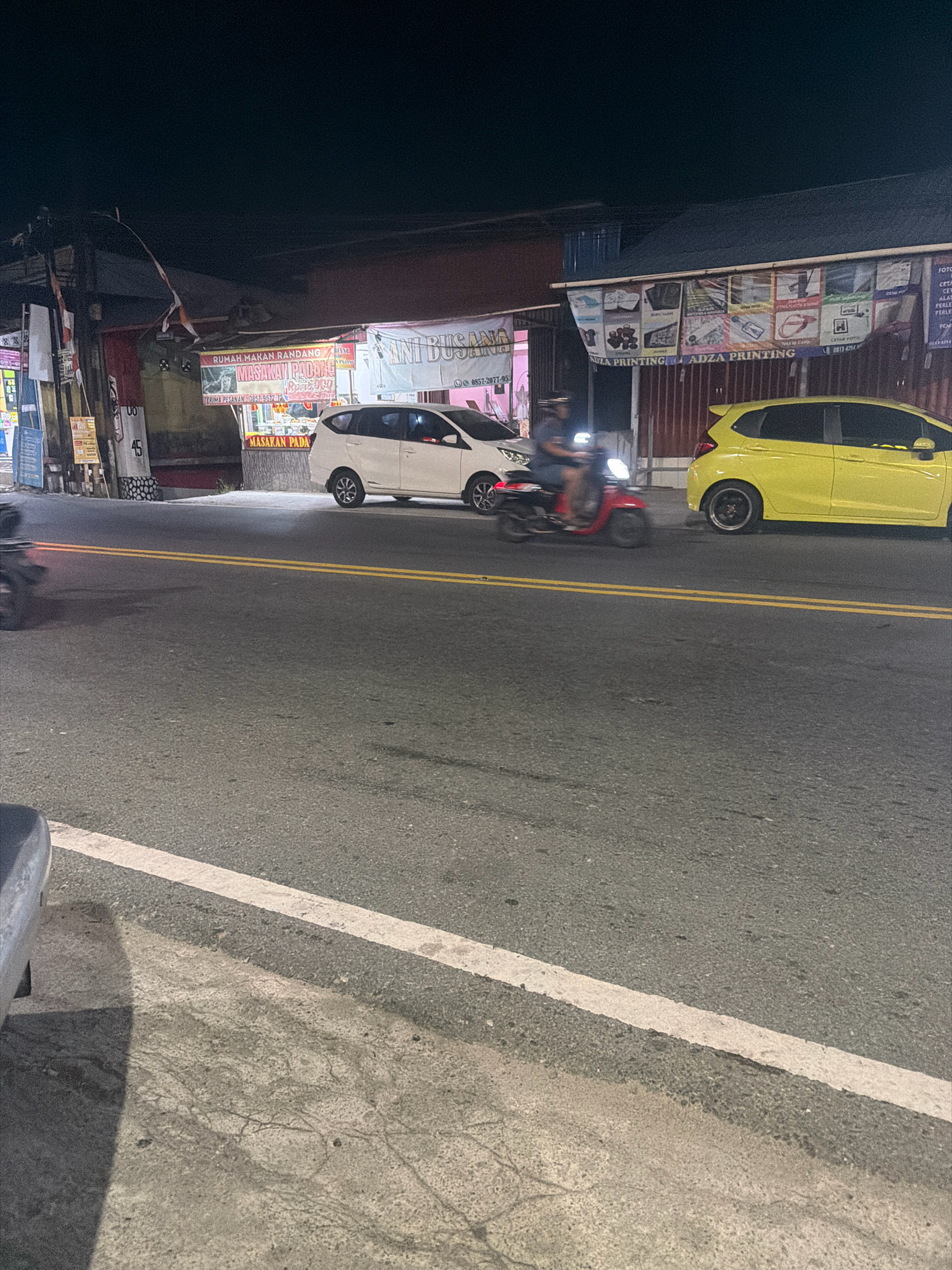 The image depicts a street scene at night in Balikpapan, Indonesia. A yellow Honda Jazz is parked on the right side of the road, while a white MPV is in motion on the left lane. A person on a red scooter, wearing a helmet and dark clothing, is riding in the left lane, heading towards the viewer. In the background, a row of buildings lines the street, displaying various signs and advertisements. One sign reads "RUMAH MAKAN RANDANG" and "MASAKAN PADANG," indicating a food establishment. Another banner, "ADZA PRINTING," suggests a printing service. The street is asphalt, marked with double yellow lines, and illuminated by ambient light from the buildings and vehicles.
