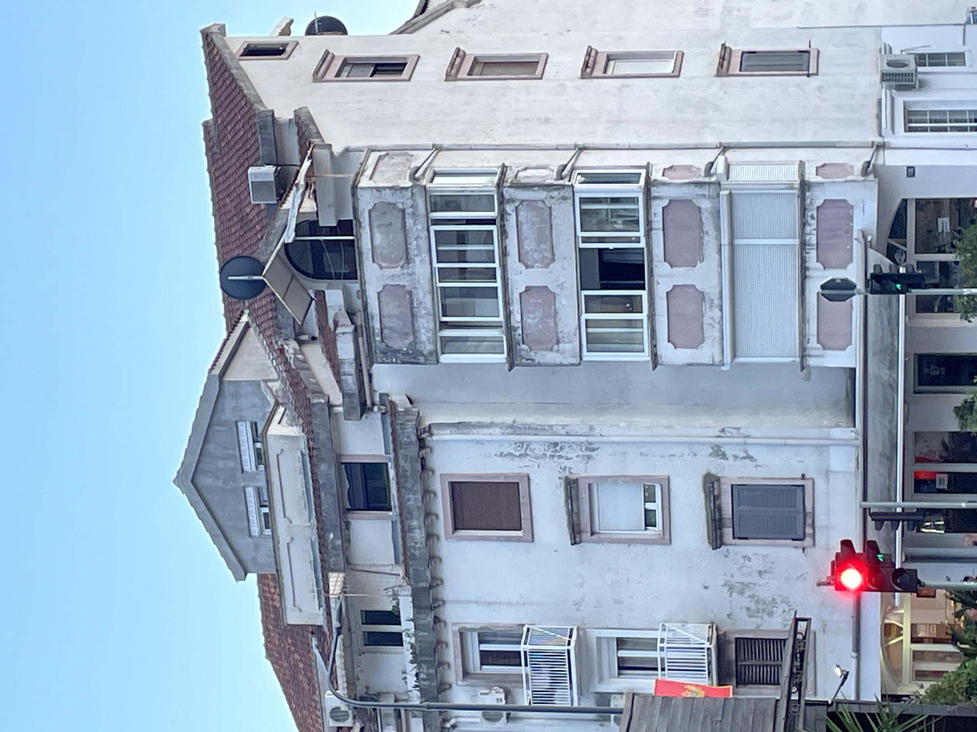 An eye-level outdoor shot of a multi-story building in Budva, Montenegro, taken in daylight. The building is white, with brown tiled roofs and several windows of various sizes. The architecture appears older, with some signs of weathering. Several of the windows are shuttered, and some have air conditioning units installed below them. A traffic light shows red on the right side of the image. The sky is a clear, pale blue.
