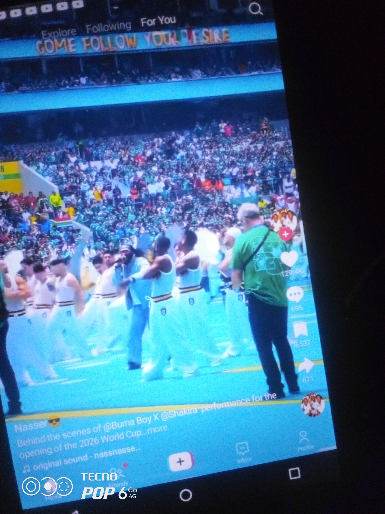 The image displays a screen recording of a TikTok video playing on a smartphone. The video shows a large crowd in a stadium, likely for a sporting or entertainment event, with a stage or performance area in the foreground. Several individuals are on the stage, some in white outfits with yellow trim, and one person in a green shirt is holding a microphone. Above the crowd, there is text displayed on what appears to be a stadium screen that reads "COME FOLLOW YOUR DESIRE."
The bottom portion of the smartphone screen shows TikTok interface elements, including a caption that reads: "Nasser Behind the scenes of @Buma Boy X @Shakira performance for the opening of the 2026 World Cup...more original sound - nassnasse..." There are also icons for likes (129.4K), comments (696), and shares (9,537). The bottom navigation bar includes icons for Home, Inbox, and Profile. The lower edge of the smartphone screen displays the "TECNO POP 6 Go 4G" logo.
Based on the information within the video's caption, specifically referencing the "2026 World Cup," this event is a preview or behind-the-scenes look at preparations for that tournament. While the TikTok caption mentions "@Buma Boy X @Shakira performance," and these artists have a history of performing at FIFA World Cup events, this particular performance is indicated to be for the *2026* World Cup. The city of Howayi, Nigeria, is not mentioned in the provided image or its context. Without additional information linking this specific event or performance to Howayi, Nigeria, it is not possible to confirm its location there.