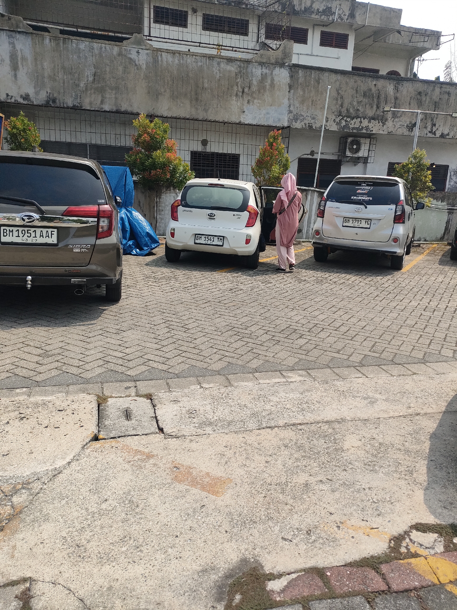 In Pekanbaru, Indonesia, a brown SUV is parked on a paved lot next to a white hatchback. A woman wearing a pink hijab and matching dress stands with her car door open, her left hand on the door frame. The SUV has a license plate reading "BM 1951 AAF". The white hatchback has a license plate reading "BM 1541 TM". A second white car with a license plate reading "BM 1791 YG" is parked behind them. The background features a building with an outdoor air conditioning unit and windows covered by bars. Small, decorative trees with red and green foliage are planted in front of the building. The ground is paved with interlocking gray bricks and concrete.