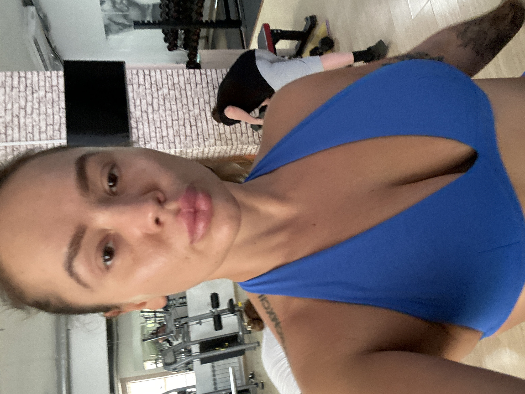 A woman with her hair pulled back in a bun, wearing a blue sports bra, is taking a selfie in a gym. Her facial expression is neutral, with her lips slightly pursed. In the background, other gym-goers can be seen, along with various exercise equipment like weight machines, dumbbells, and a weight bench. The gym appears to be indoors, with brick-patterned walls and large mirrors reflecting the space.