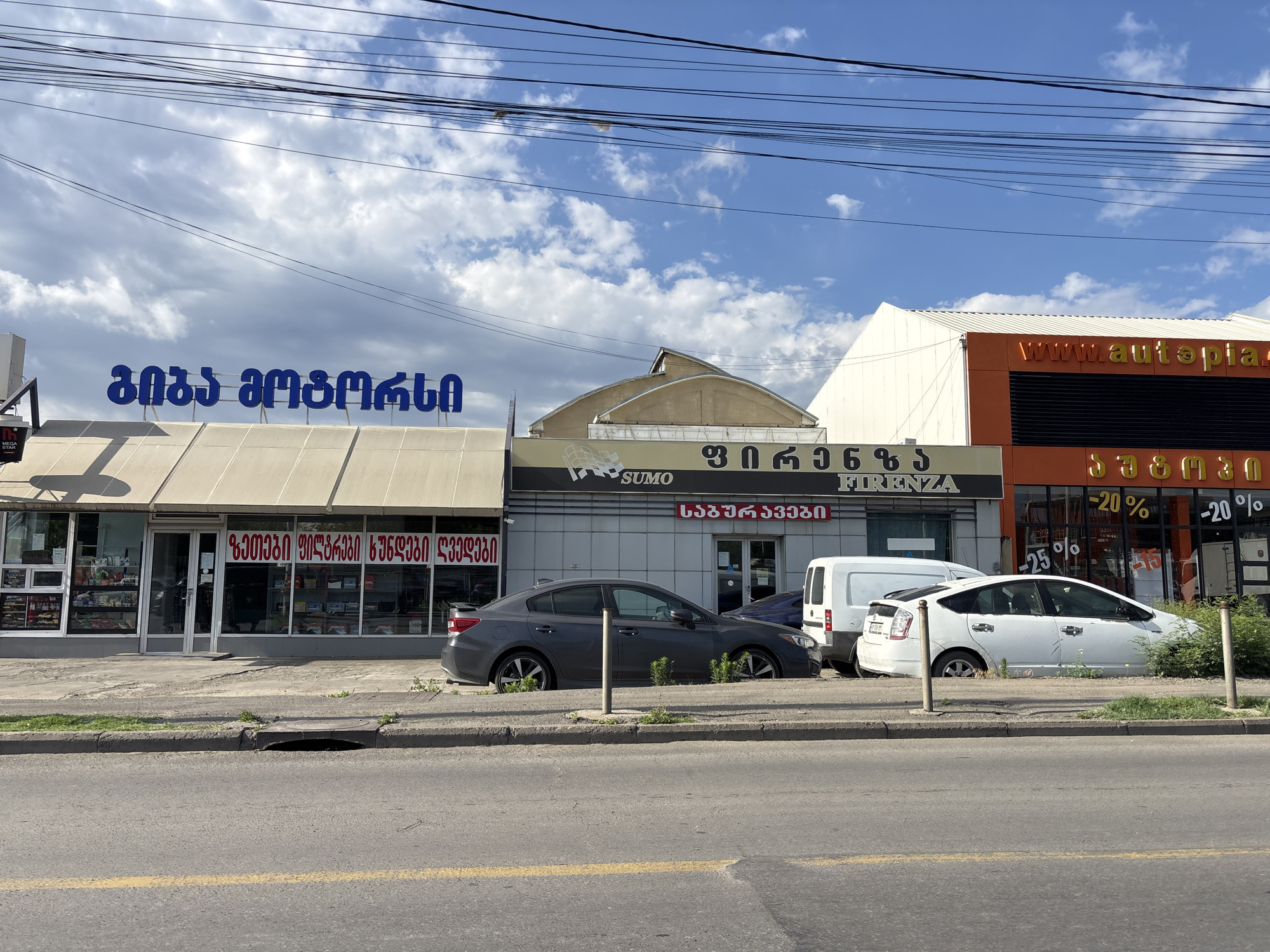 The image shows a street scene in Tbilisi, Georgia, with a row of shops along the side of the road. The shops have signs with text in Georgian script. The leftmost shop has a blue sign with white lettering. The middle shop has a dark grey facade with a sign that reads "SUMO FIRENZA". The rightmost shop has an orange facade with a sign that reads "www.autopla" and other text in yellow lettering. In front of the shops, there are several cars parked along the curb, including a dark grey sedan and a white hybrid vehicle. The street is paved and there is a yellow line marking the lane. There are power lines visible overhead. The sky is blue with scattered clouds. The environment appears to be an urban commercial area.
