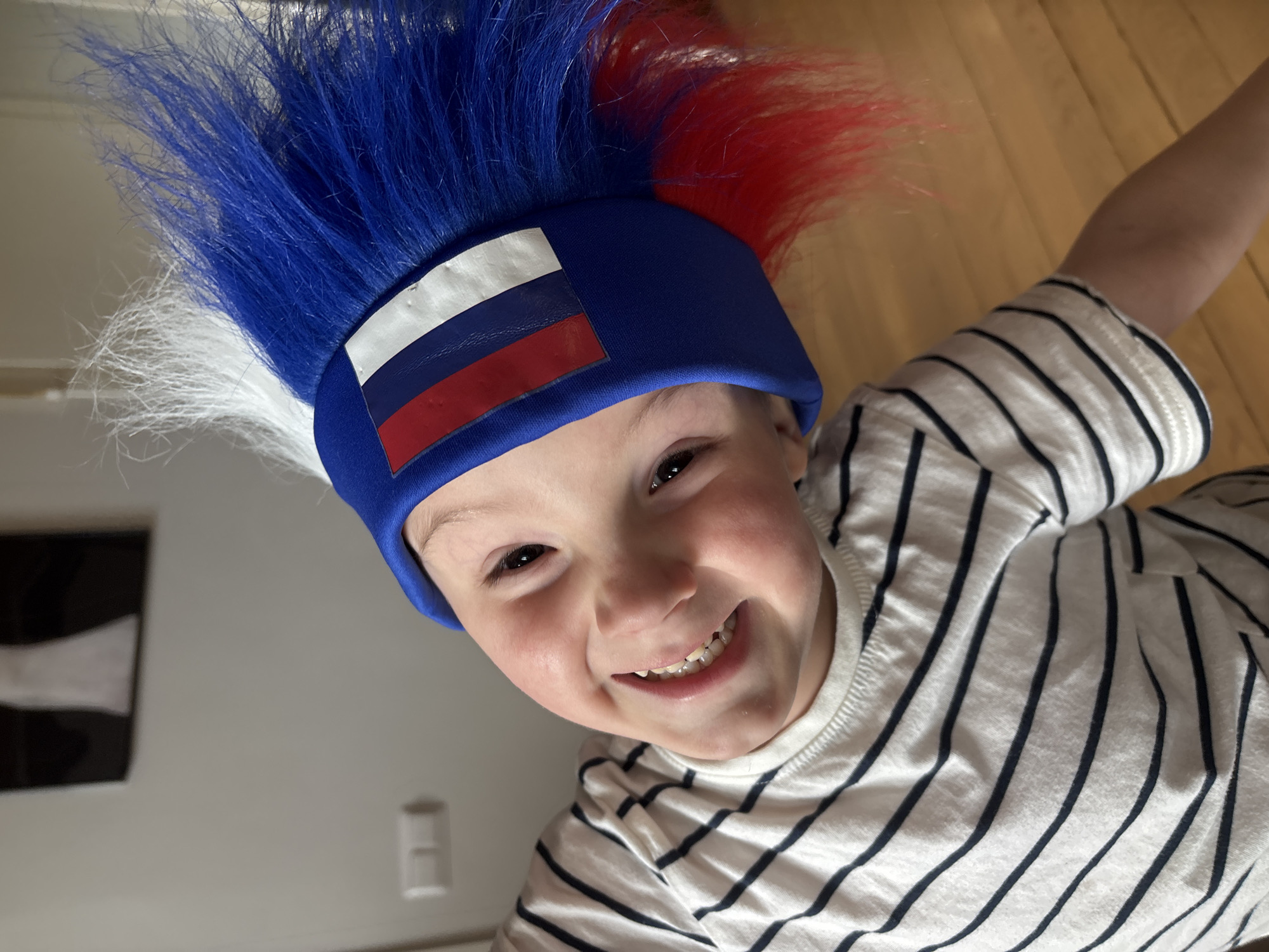 A young child is photographed from above, smiling broadly with teeth showing. The child is wearing a hat adorned with the Russian flag and an attached "hair" made up of blue, white, and red fibers. The child is wearing a white shirt with thin navy blue stripes. The background includes a light-colored wall with a dark rectangular object (possibly a picture or a painting) and light-colored wooden flooring. There is no explicit evidence indicating the location is Chaikovsky, Russia.