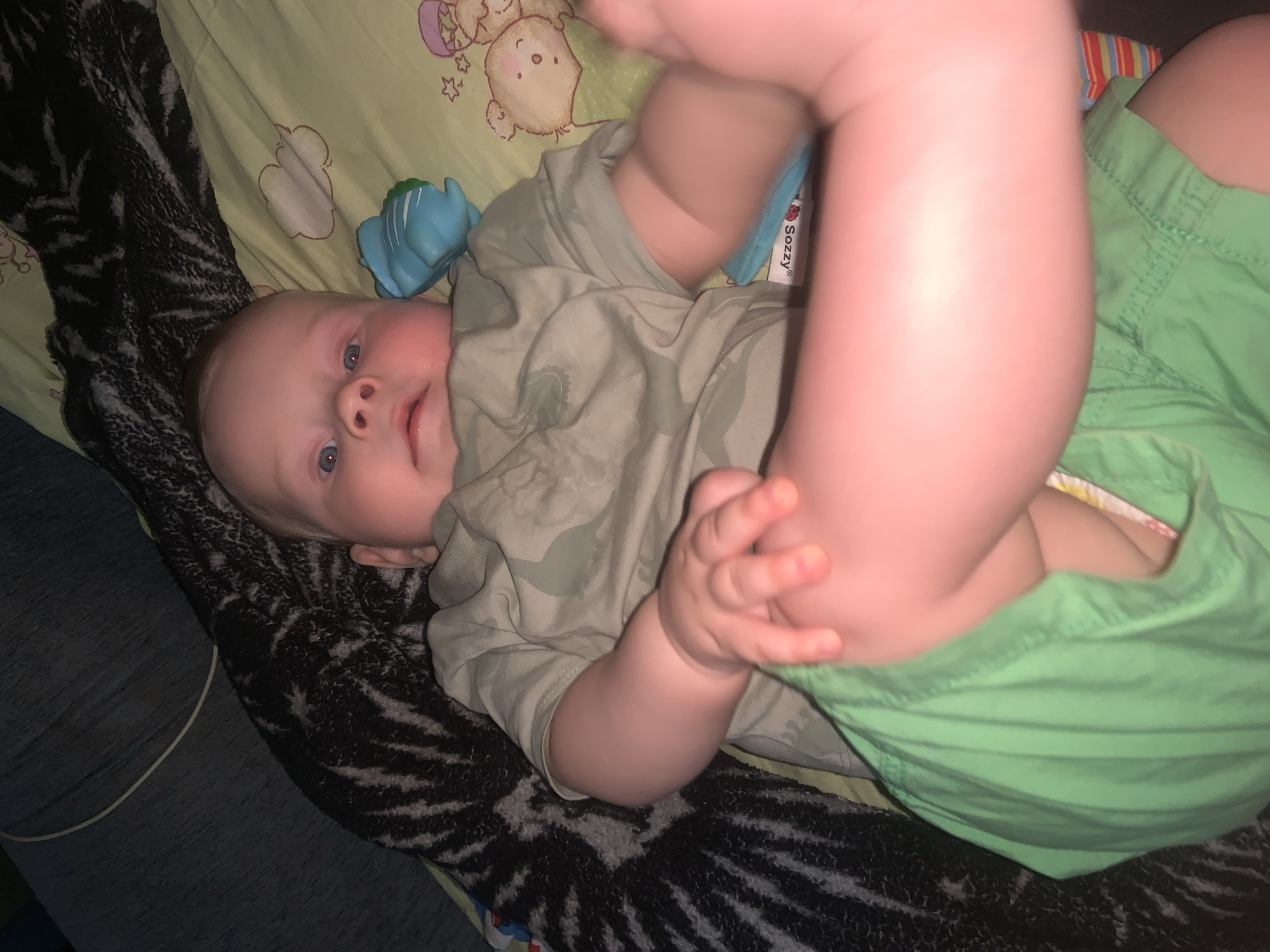A baby with blue eyes and light hair is lying on their back on a light green blanket with teddy bear prints. The baby is wearing a light green t-shirt with a camouflage pattern and green shorts. A blue toy is next to the baby's head. The background includes a dark blanket with a black and white striped pattern, and a portion of a dark blue surface. The lighting suggests an indoor setting, possibly at night. The location is unknown.